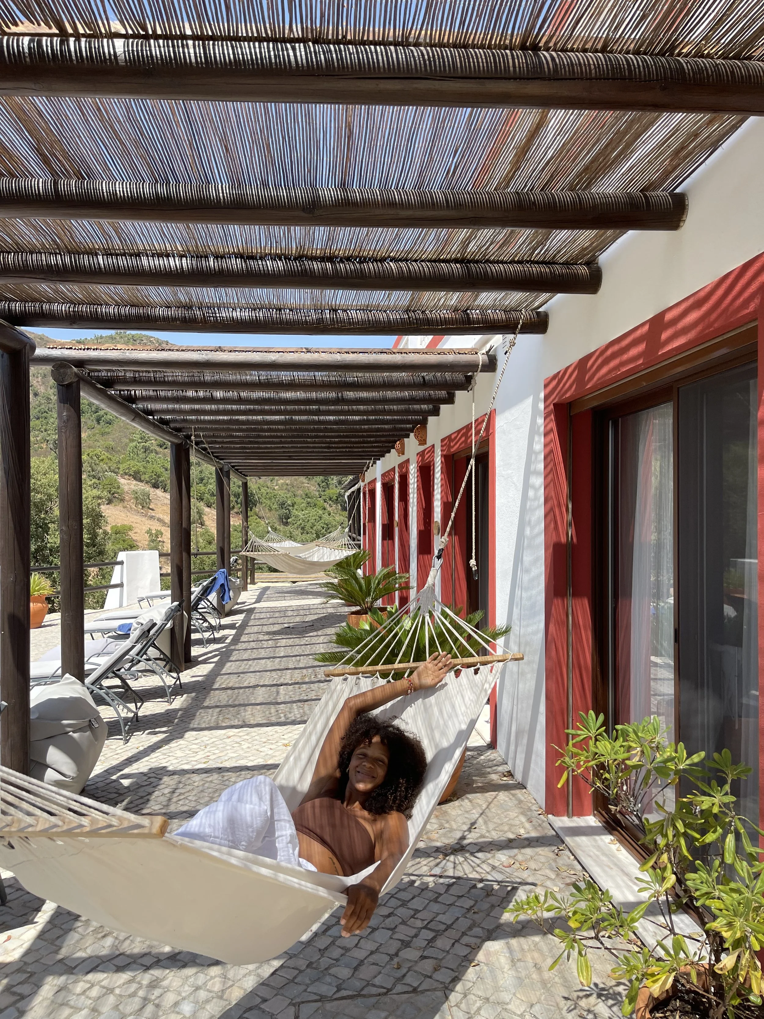 Woman relaxing on a hammock on a sunny patio with lounge chairs, potted plants, and a scenic hillside in the background, on a yoga retreat in Portugal.