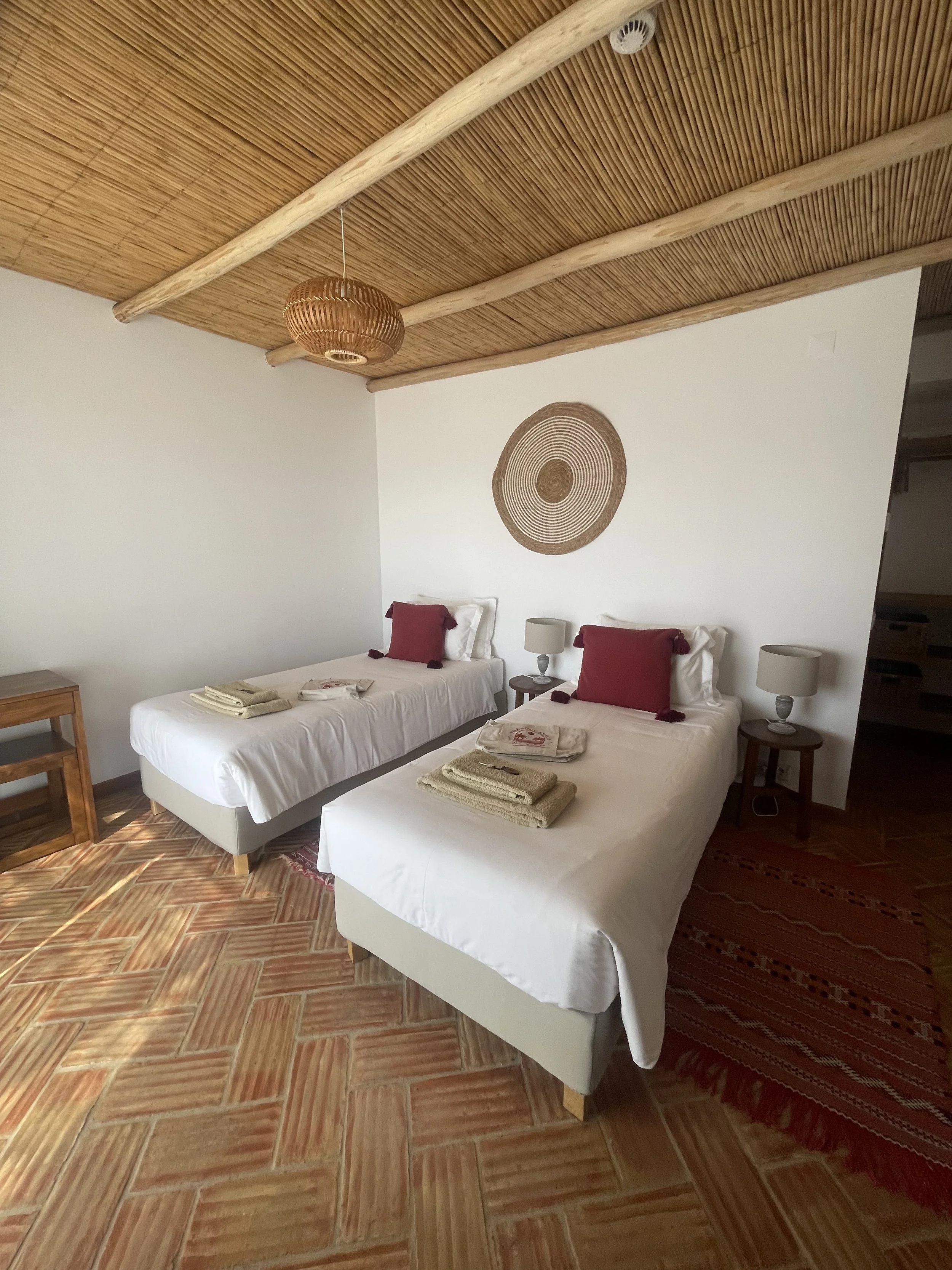 A bedroom with two single beds, each with white linens, red pillows, and folded towels on top. There are two nightstands with lamps between the beds. A round woven wall decoration is above the beds. The room features wooden furniture, a wooden ceilin