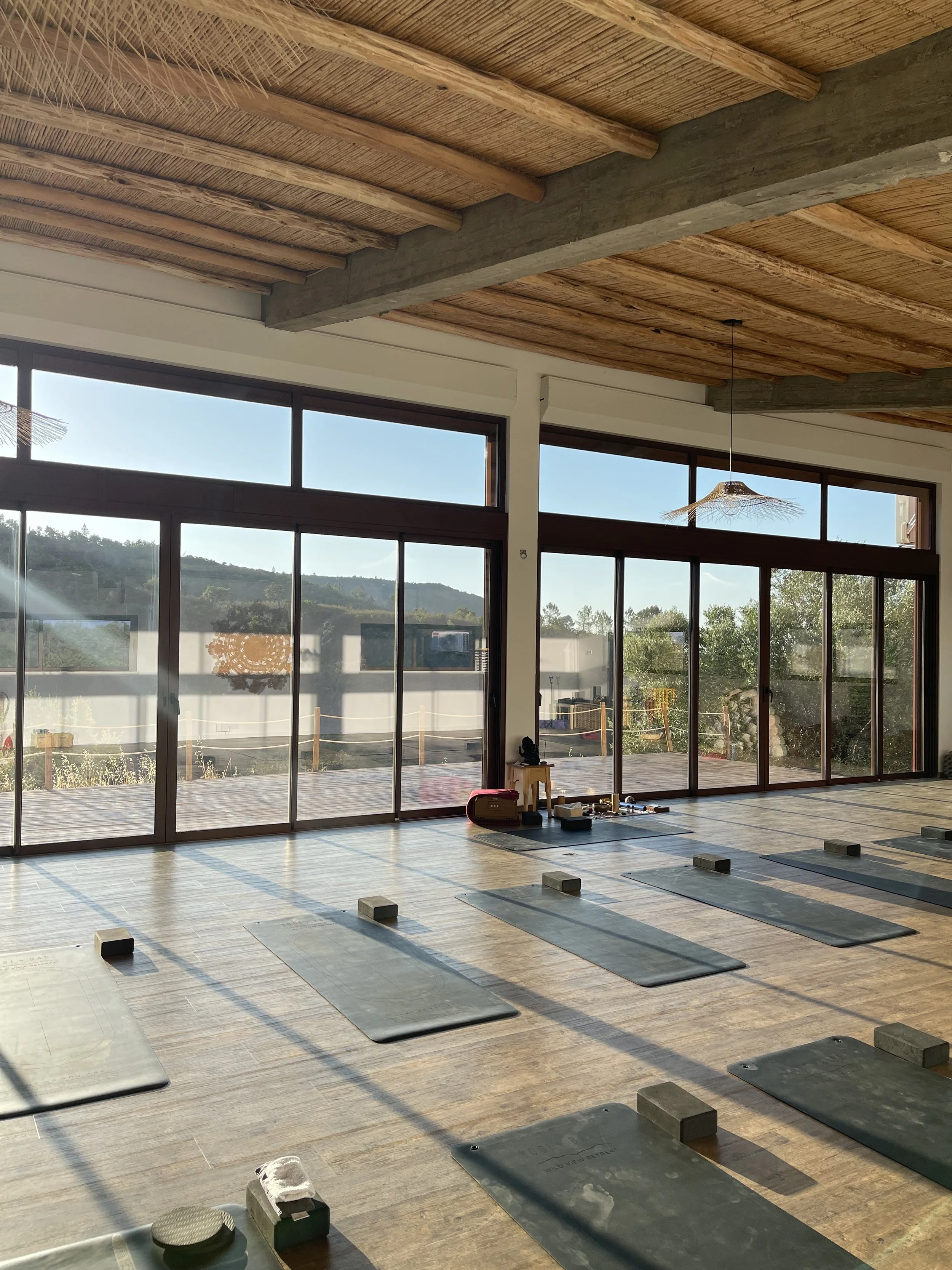 An empty yoga studio with large glass sliding doors, yoga mats on the wooden floor, and a view of trees and mountains outside. The ceiling has wooden beams and a hanging light fixture, on a yoga retreat in Portugal.