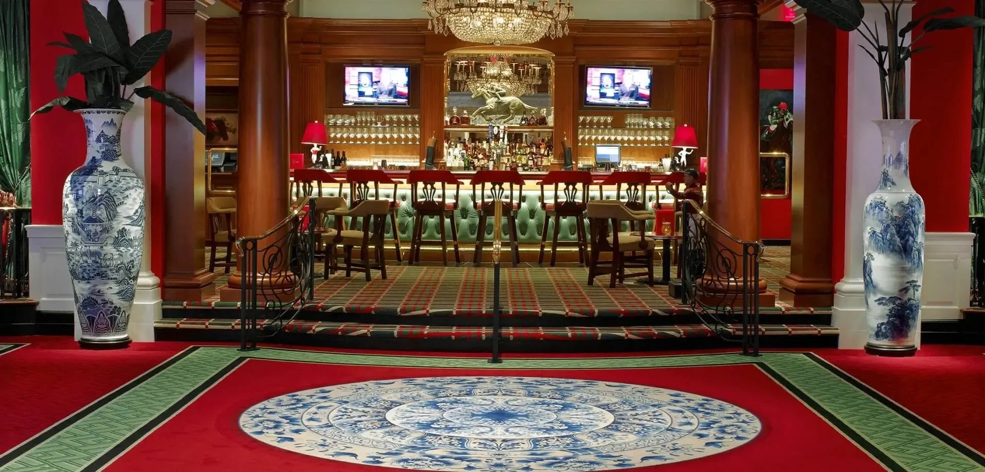 Inside a luxurious bar with a red and green color scheme, featuring two large blue-and-white porcelain vases with plants, a red carpet, and a bar counter with high chairs, lamps, and a mounted horse sculpture on the back wall.