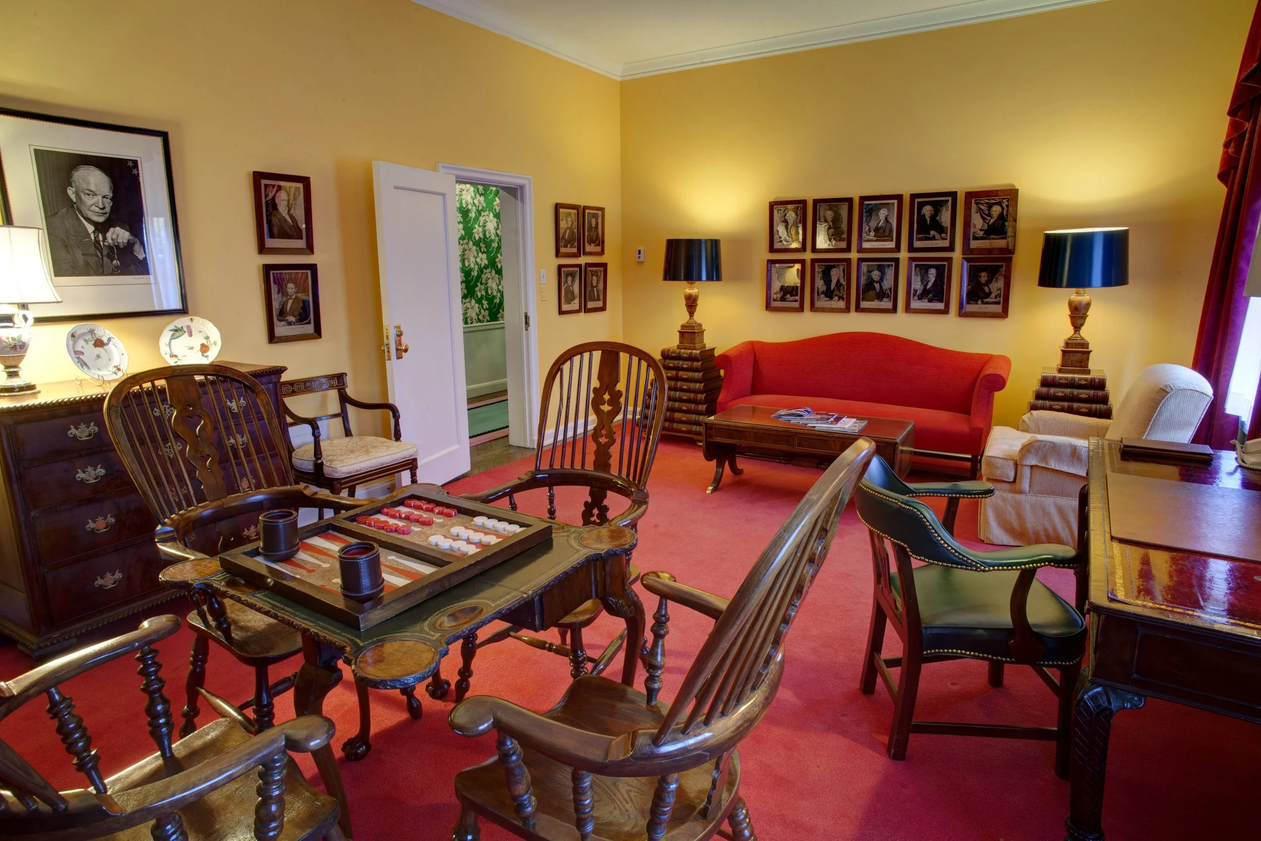 Living room with yellow walls, red carpet, and antique furniture, including a wooden dining table with chairs, a red sofa, an armchair, coffee tables, lamps, and framed photographs on the wall.