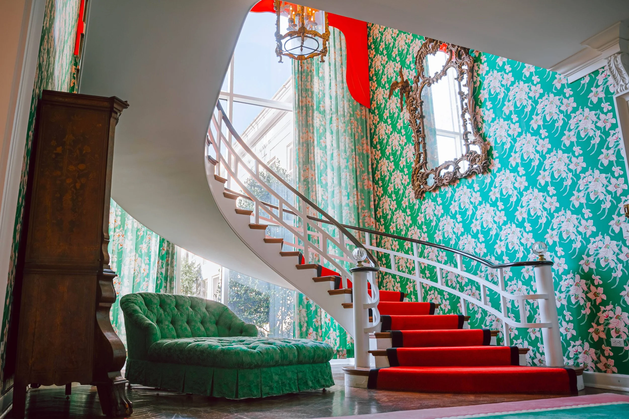 Interior of a colorful home with a green tufted sofa, a spiral staircase with red carpeting, floral wallpaper, big windows with curtains, an ornate mirror, and a chandelier.