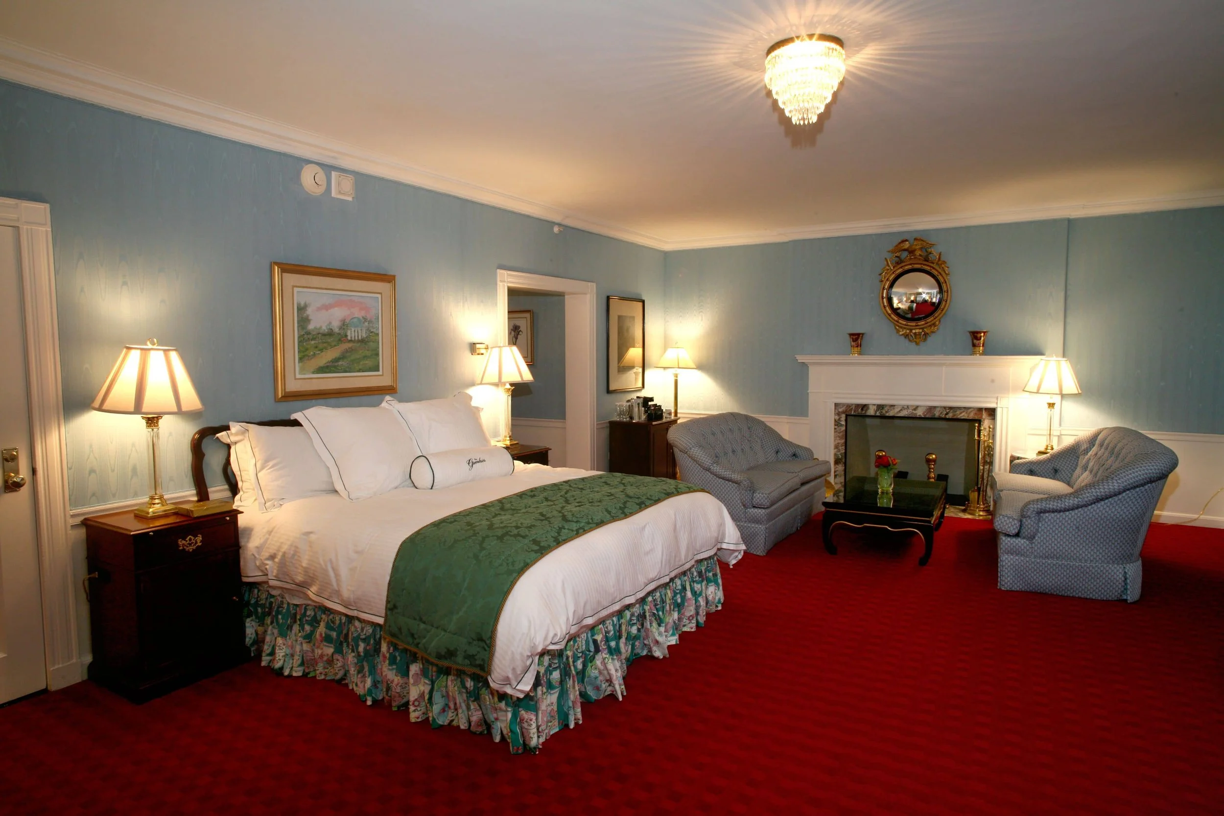 A spacious hotel room with a large bed, two nightstands with lamps, a sitting area with two blue armchairs and a small sofa, and a fireplace with a mirror above it. The room features blue walls, a red carpet, and decorative artwork.