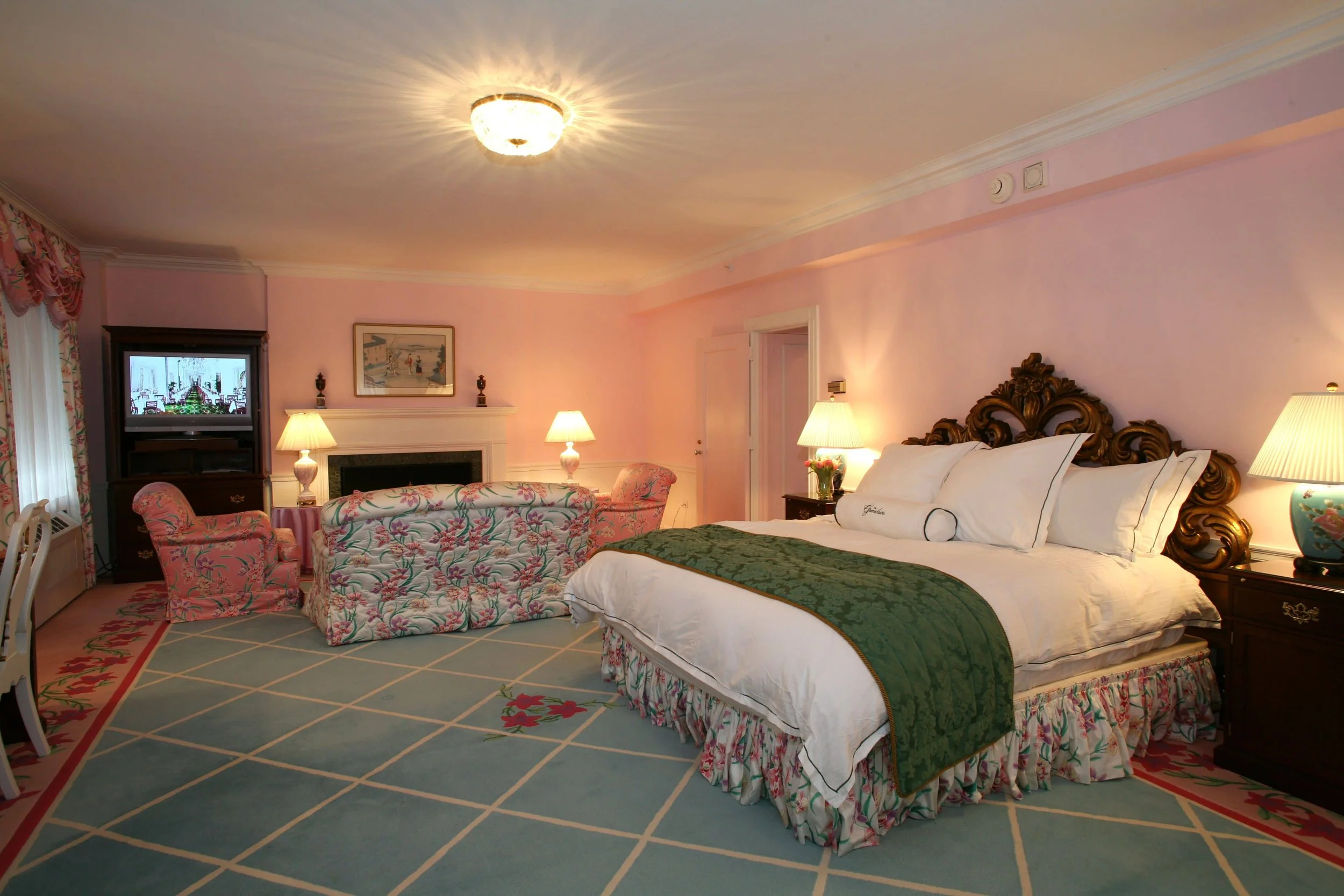 A luxurious hotel room with a large ornate wooden headboard, white bedding with a green throw, bedside tables with lamps, a sitting area with floral upholstered chairs and a sofa, a fireplace, and a TV in a wooden cabinet, all in a pastel pink color
