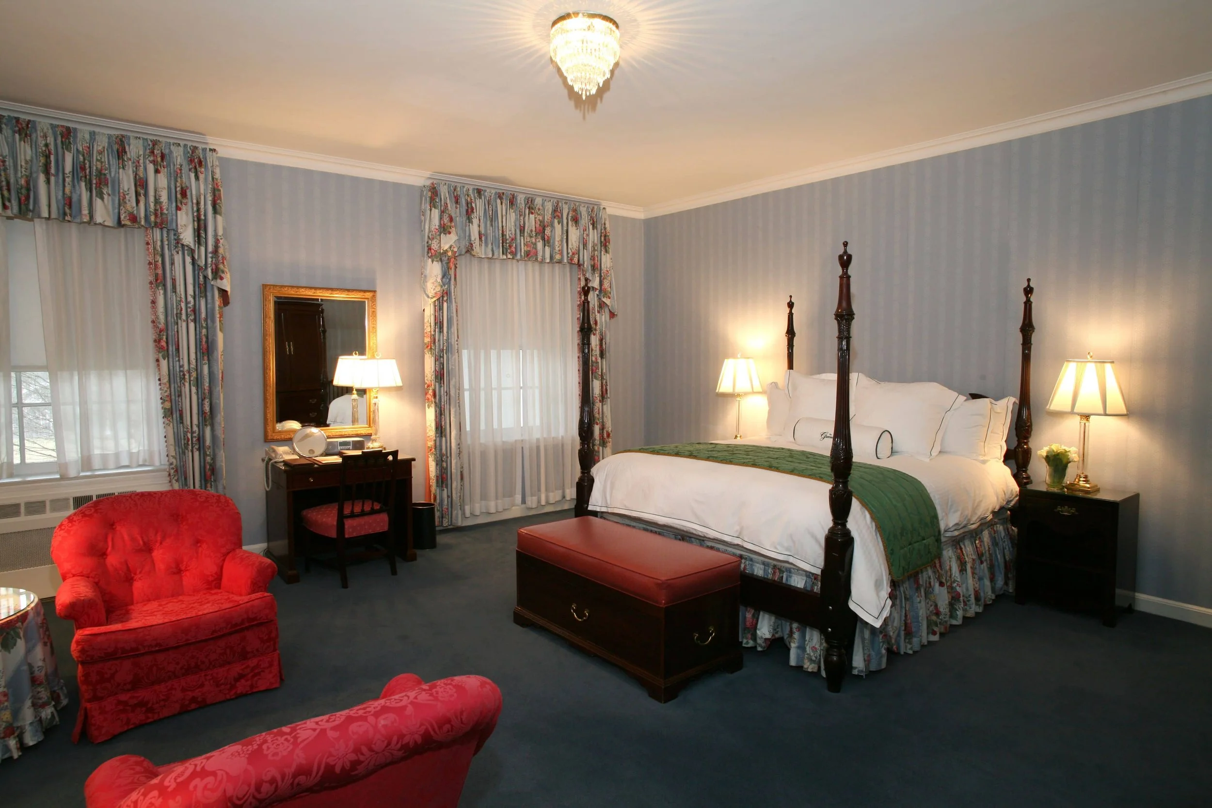 A cozy hotel room with a four-poster bed, nightstands with lamps, a red armchair, a small table, floral curtains, and a window with sheer curtains.