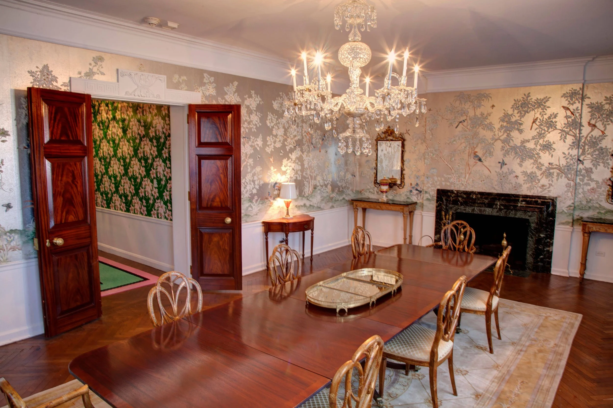 Elegant dining room with a large wooden table and eight matching chairs. A crystal chandelier hangs from the ceiling, and a decorative fireplace is on one wall. The room features floral wallpaper, a mirror, and small side tables with lamps.