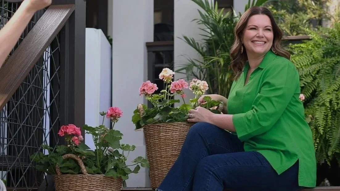 A woman with brown hair wearing a bright green shirt and dark blue jeans, smiling and sitting on outdoor steps with potted pink and white flowers.