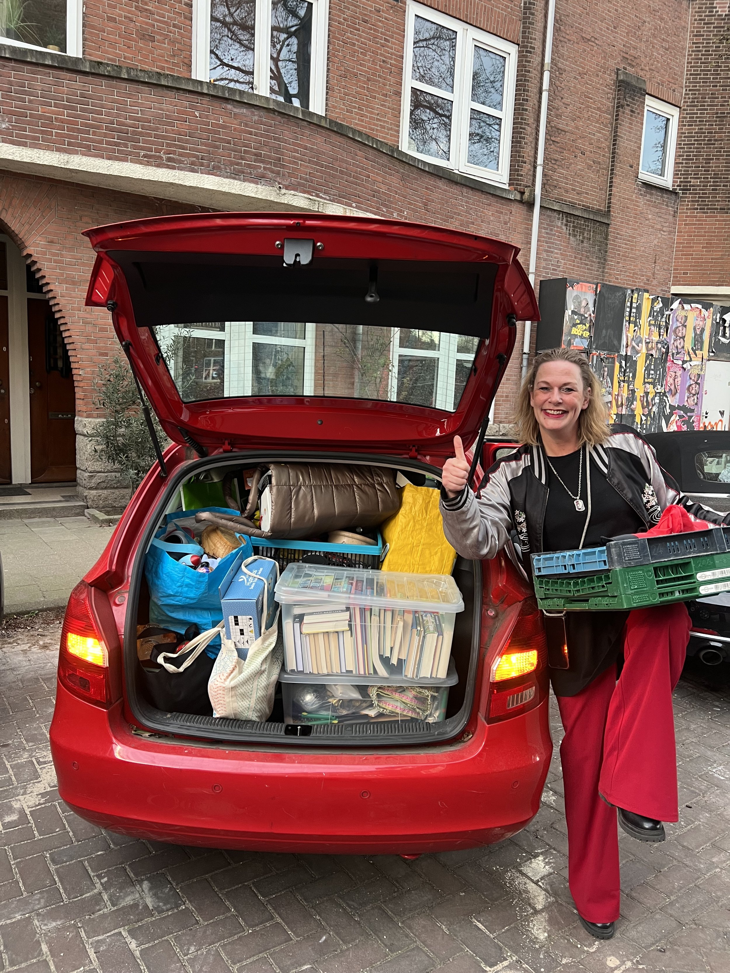 Vrouw met glimlach en duim omhoog naast de achterklep van een rode auto, volgeladen met dozen en tassen.