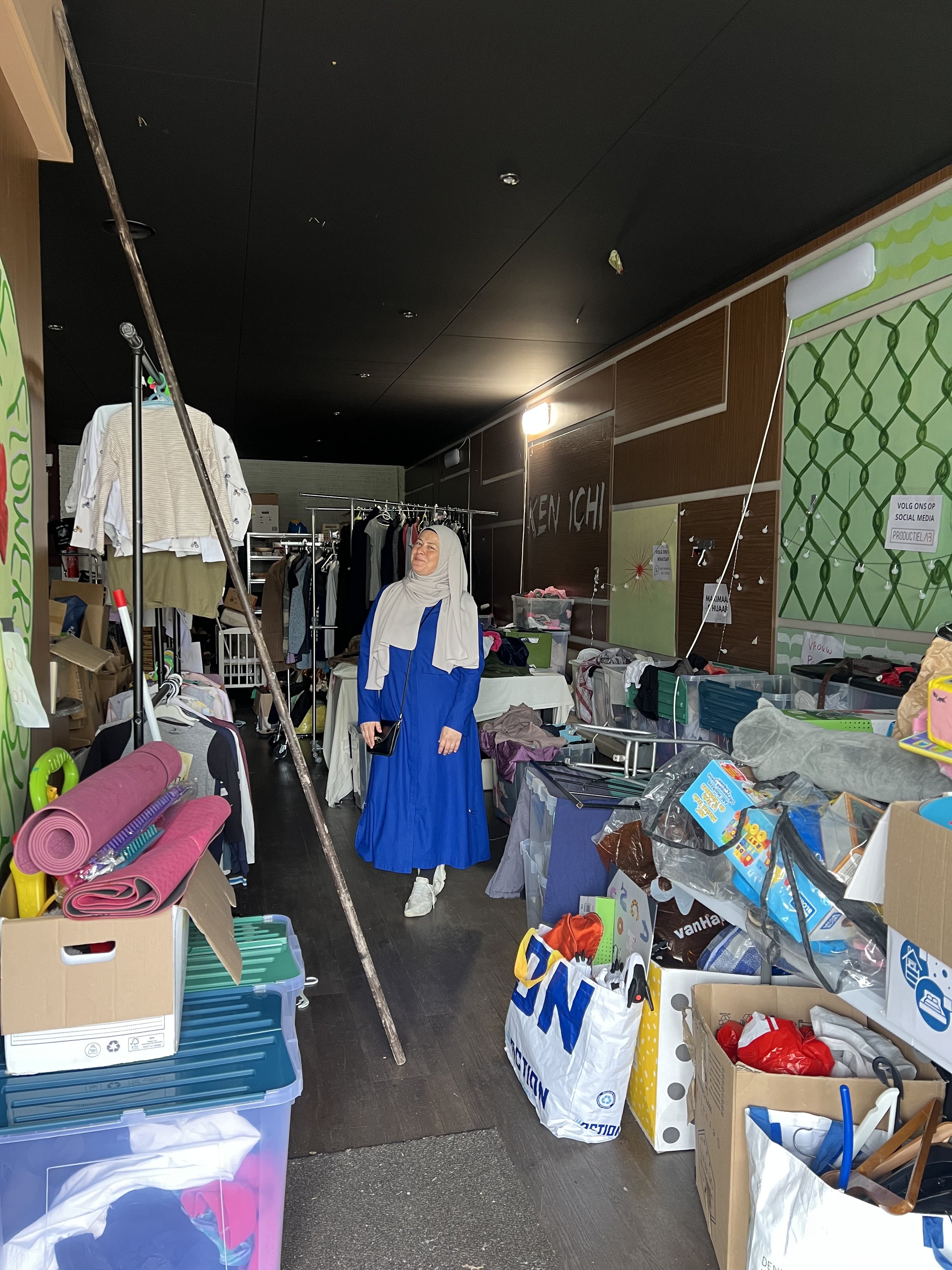 Binnen een winkel of op een markt vol met meubels, kleding en andere tweedehands artikelen. Een vrouw in een blauwe jurk en een hoofddoek staat tussen de spullen.