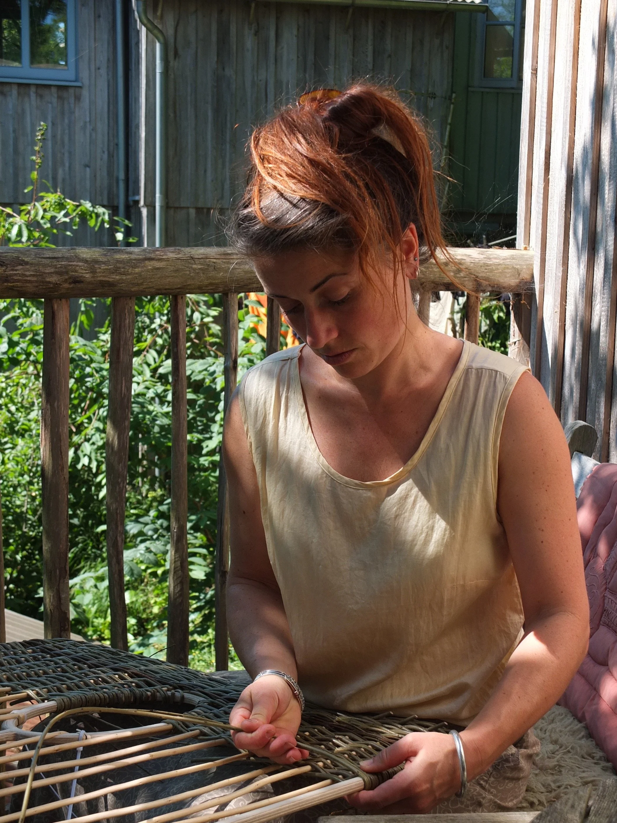 A woman with reddish-brown hair in a ponytail, wearing a beige sleeveless top, is weaving or working with natural materials outdoors on a wooden deck surrounded by greenery and a wooden fence.