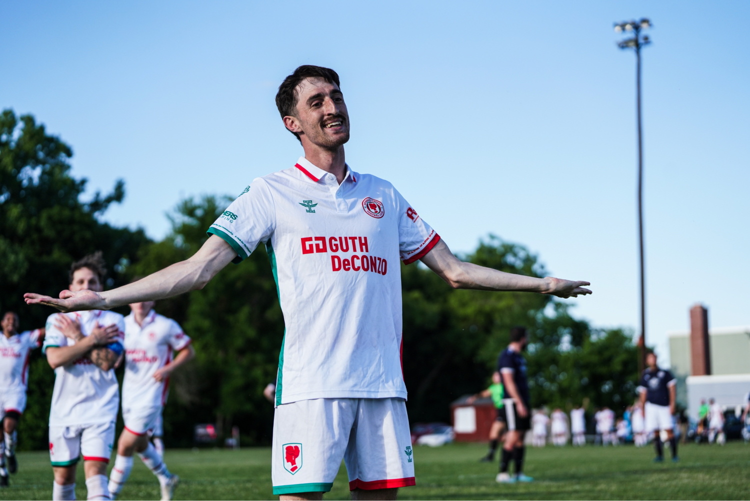 A soccer player in a white jersey with red and green accents stands on a field with arms outstretched, smiling after a game. Other players and officials are visible in the background.