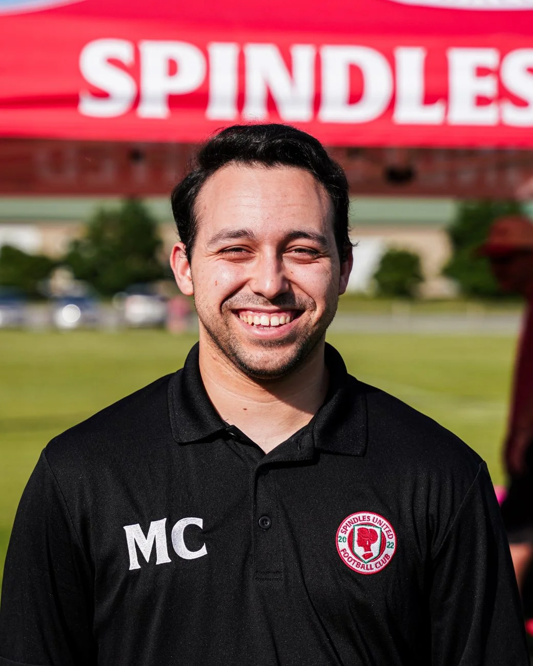 Smiling man with dark hair wearing a black shirt with initials 'MC' and a 'Spindles United Football Club' logo, standing outdoors with a red 'SPINDLES' sign in the background.