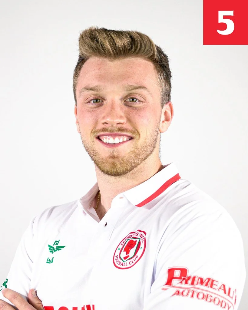 Young man with blond hair and a beard smiling, wearing a white football jersey with red and green details, and a logo for a football club.