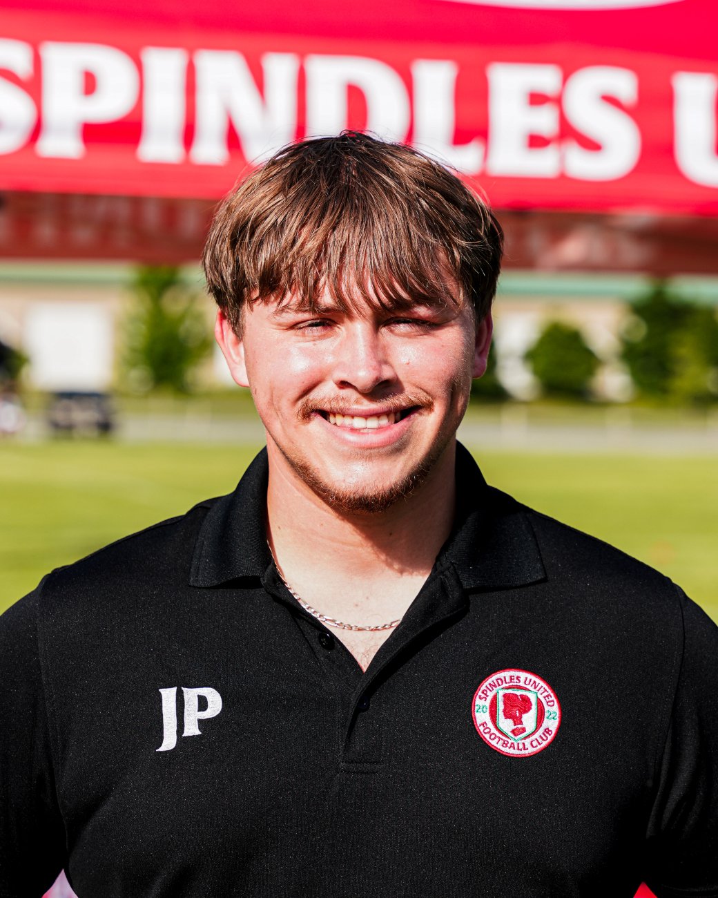A young man with brown hair smiling, wearing a black Spindles United Football Club polo shirt with the initials JP and a logo, standing outside with a blurred background of trees and a red banner.