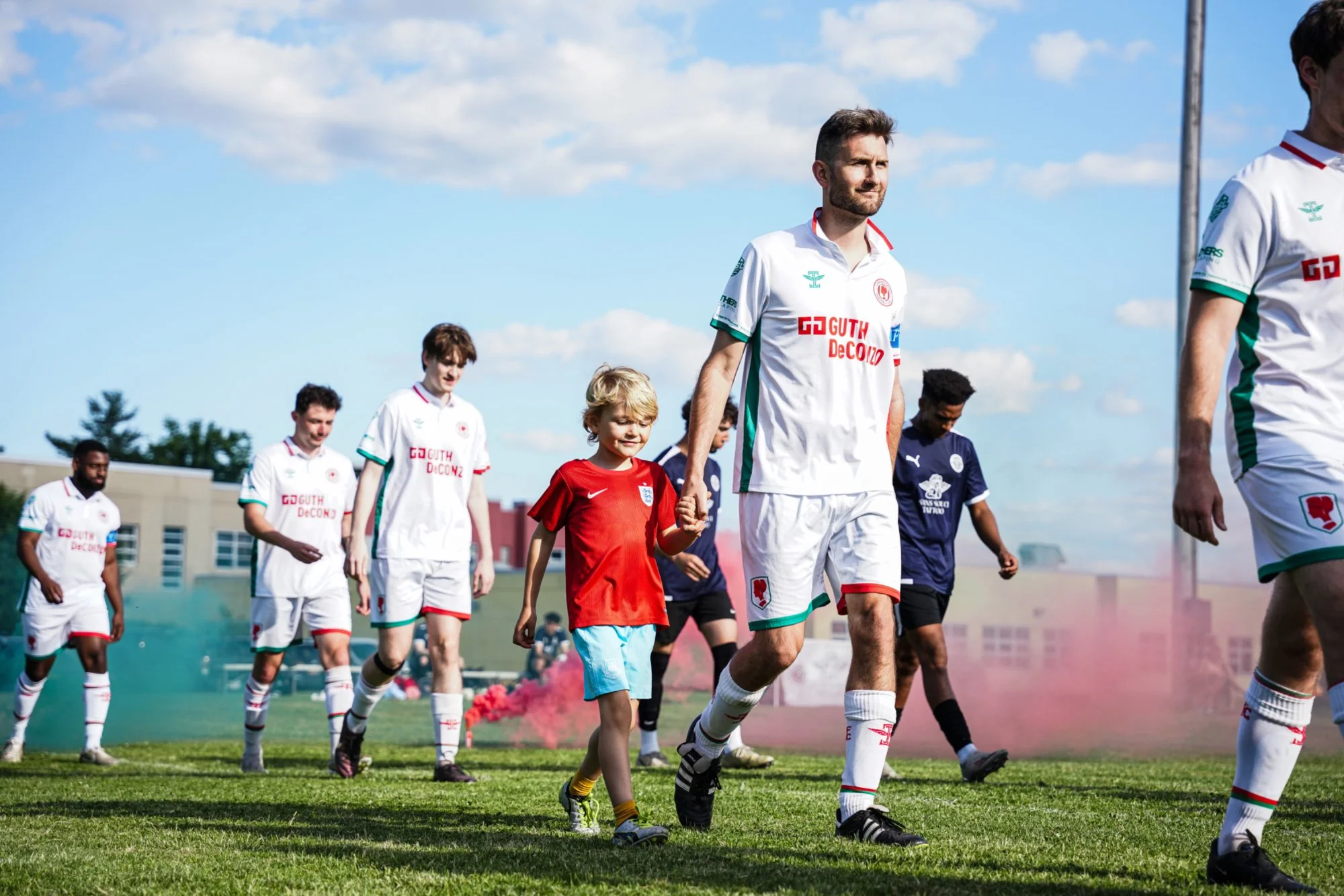 Soccer players walking onto the field, including children and adults, with some smoke effects in the background, on a sunny day with clouds in the sky.