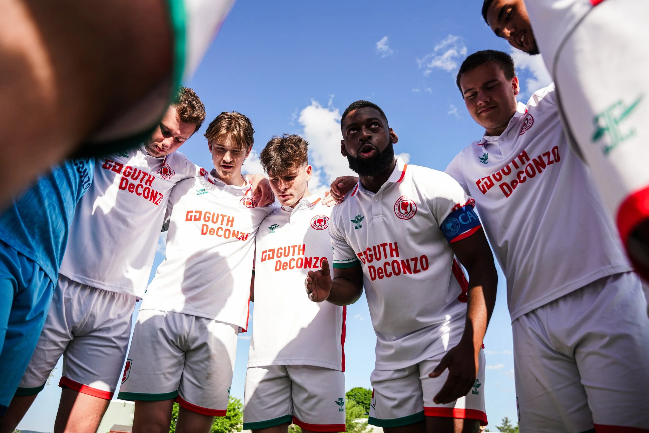 Group of soccer players in a huddle, wearing white jerseys with red and green accents, standing outdoors under a blue sky with some clouds.