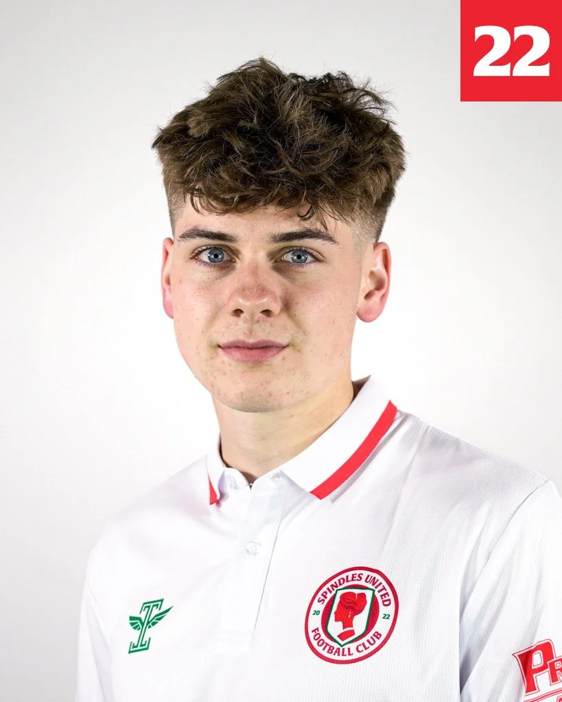 Young man with curly brown hair and blue eyes wearing a white football jersey with red and green logo of Spindles United Football Club and the number 22 in red at the top right corner.