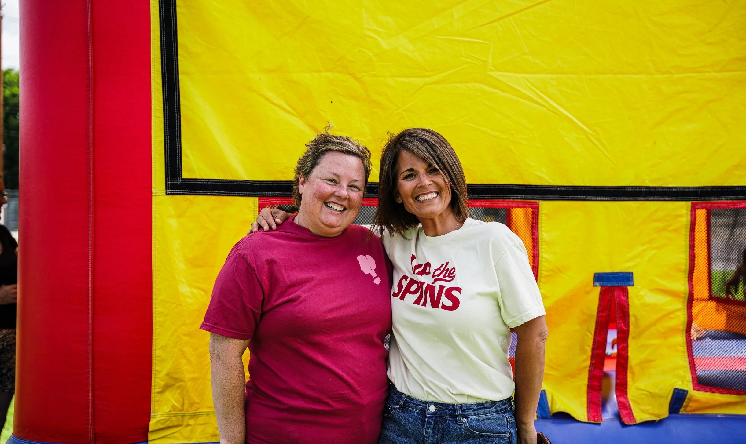 Two women smiling and standing close together in front of a yellow inflatable structure, one wearing a maroon shirt and the other a white shirt with red text.