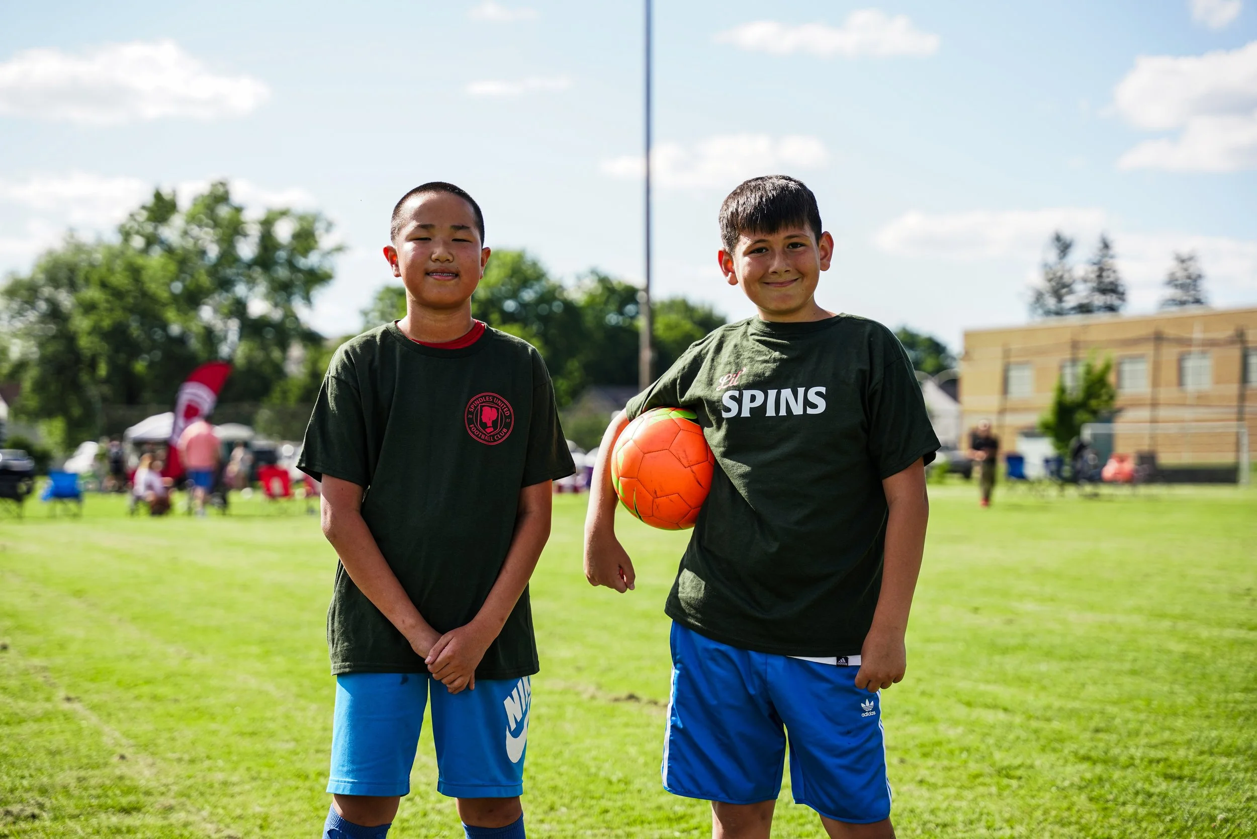 Two young boys standing on a soccer field, smiling. One boy is holding a green and orange soccer ball, wearing a black shirt with red and white text, and blue shorts. The other boy is wearing a black shirt with a red logo and blue shorts. In the back