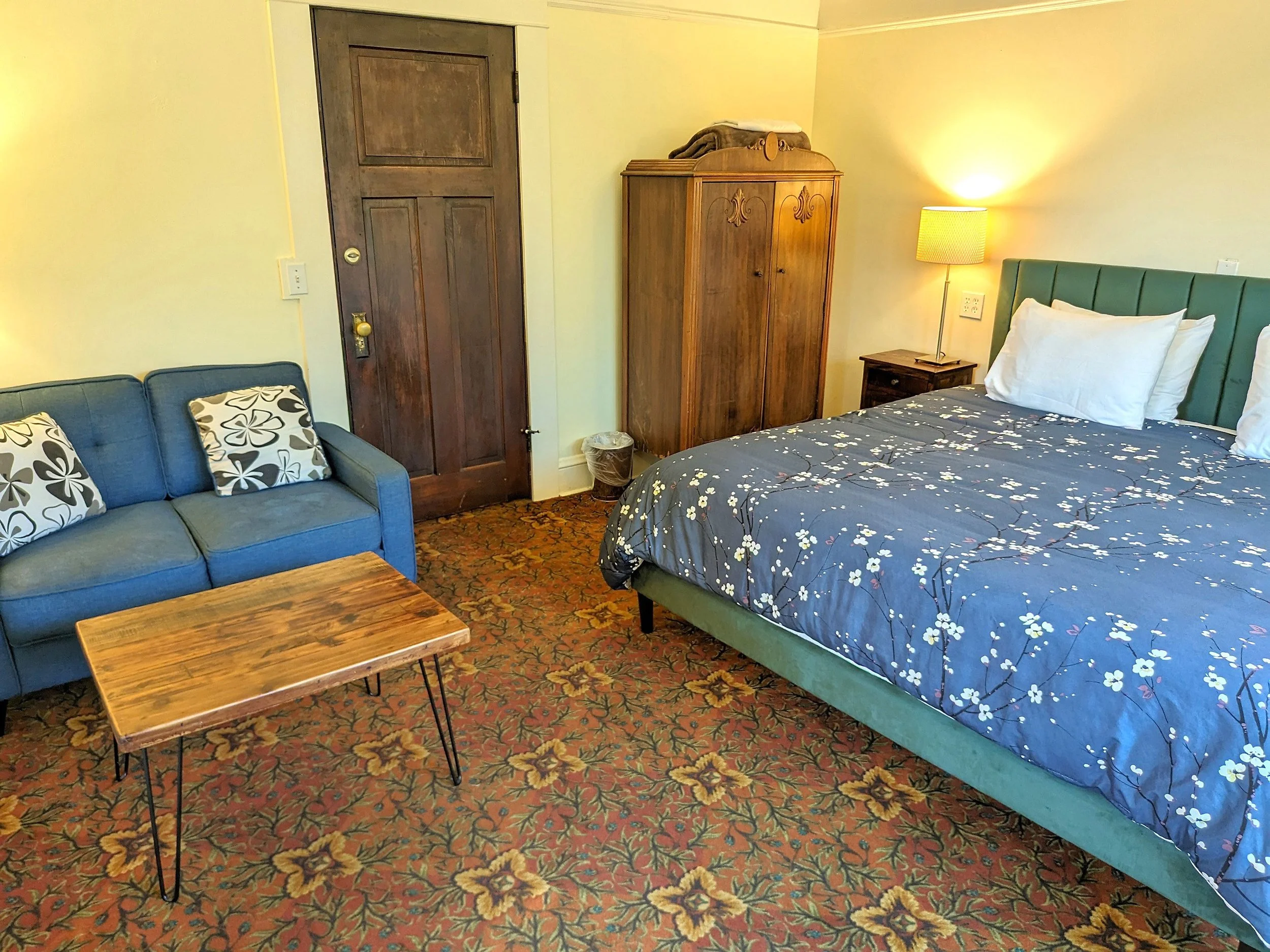 A cozy hotel room with a green bed, white pillows, and floral bedding, a wooden wardrobe, a bedside table with a lamp, a small wooden coffee table, a blue sofa with floral pillows, and a patterned carpet with floral designs.