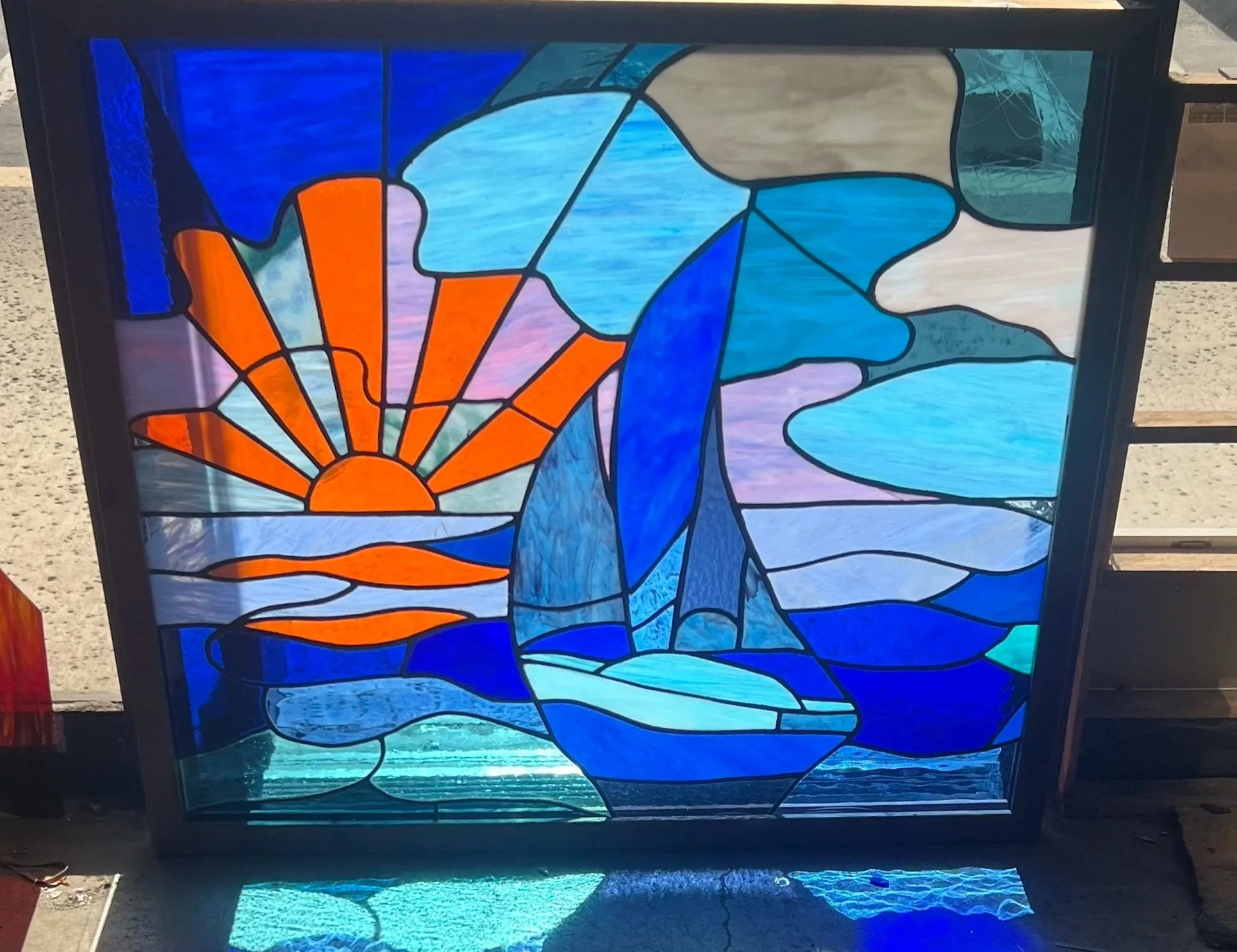 Stained glass window repair showing a landscape with a setting sun, blue sky, clouds, and flowing water.