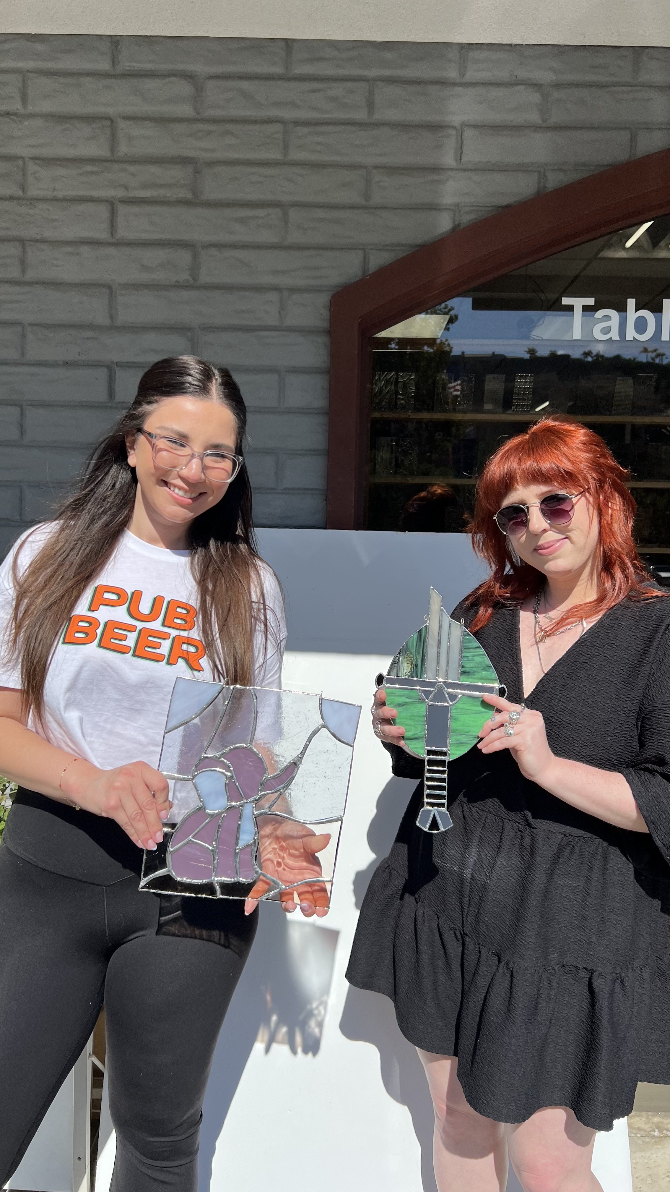 Two women standing outside, each holding a stained glass artwork. The woman on the left is wearing glasses, a white T-shirt that says 'PUB BEEER' in orange and green letters, and black pants. The woman on the right has red hair, glasses, and is wearing a black textured dress. They are smiling and posing in front of a gray brick wall and a window that reflects trees and a blue sky.