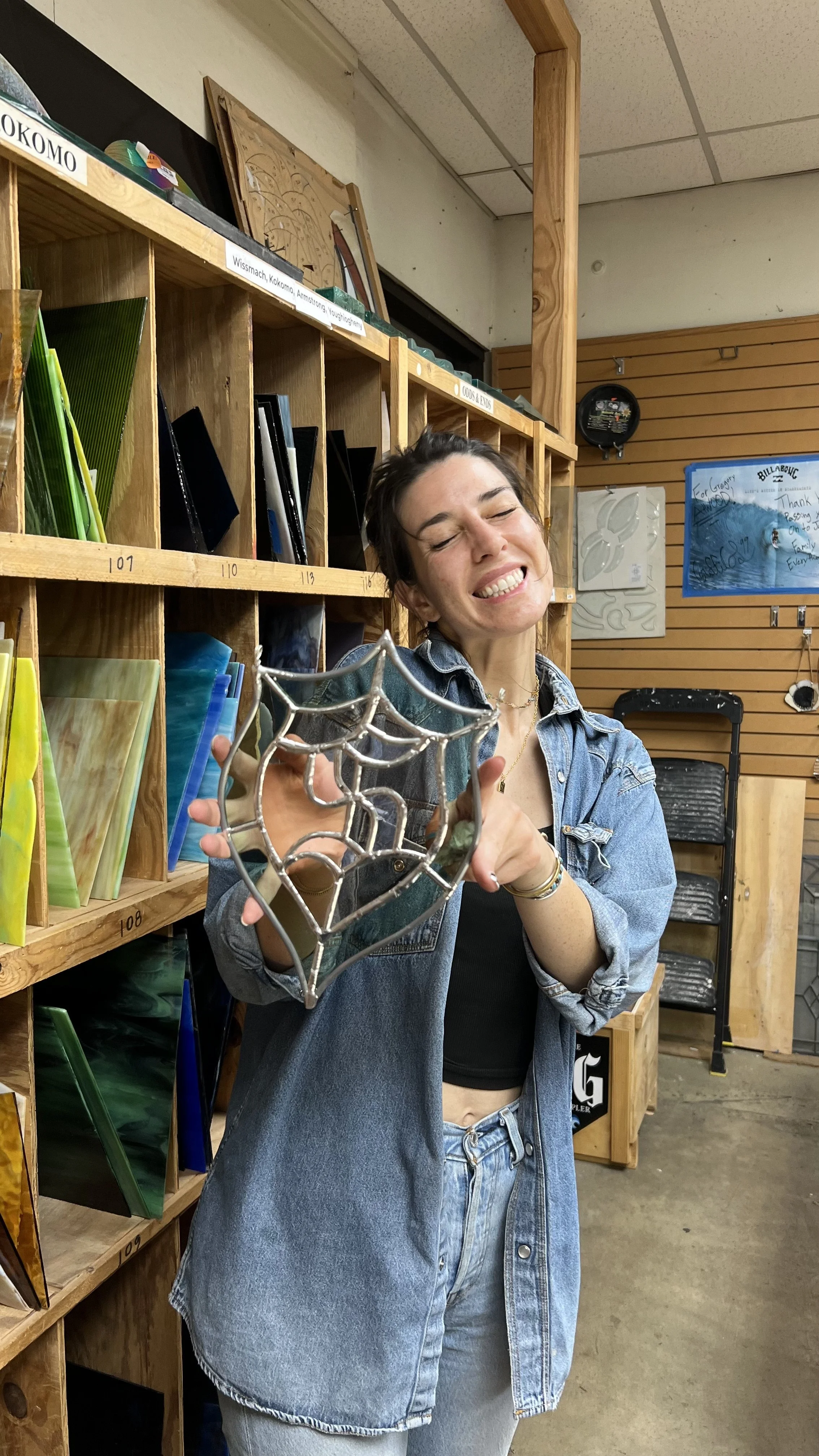 A woman with a big smile holding a small metallic spider web decoration in a store with wooden shelves filled with art supplies and decorative items.