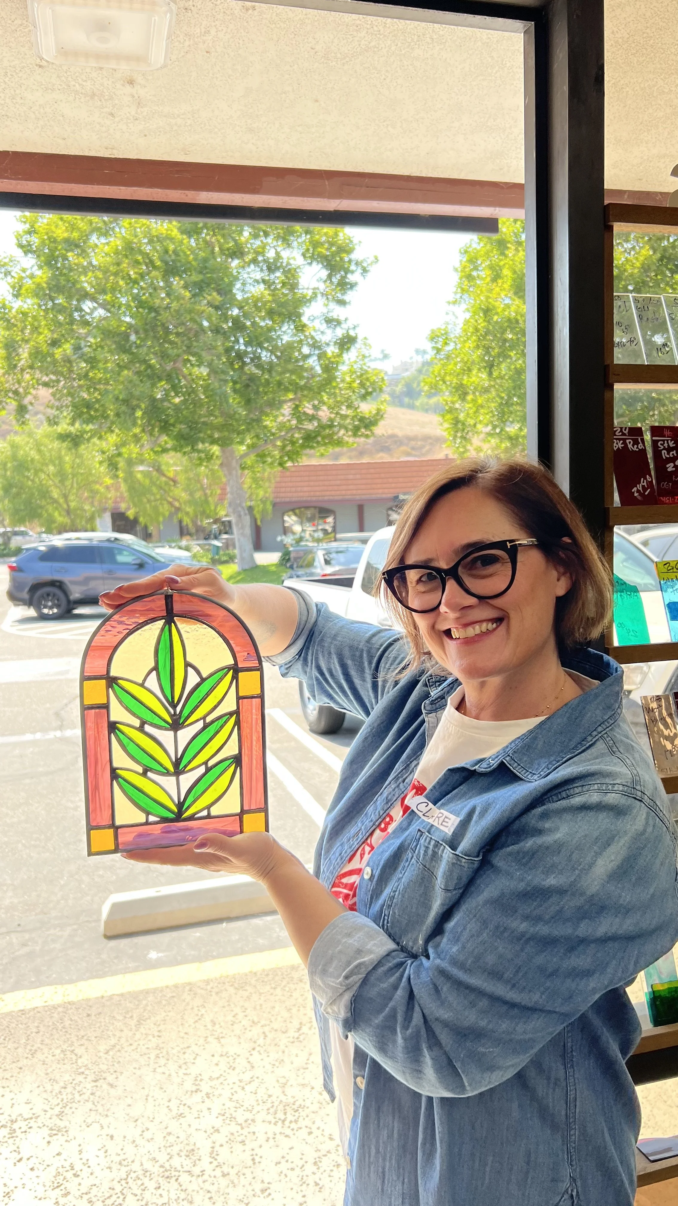A woman smiling and wearing glasses and a denim jacket, holding a colorful stained glass art piece with a leaf design, inside a store with a large window showing trees and parked cars outside.