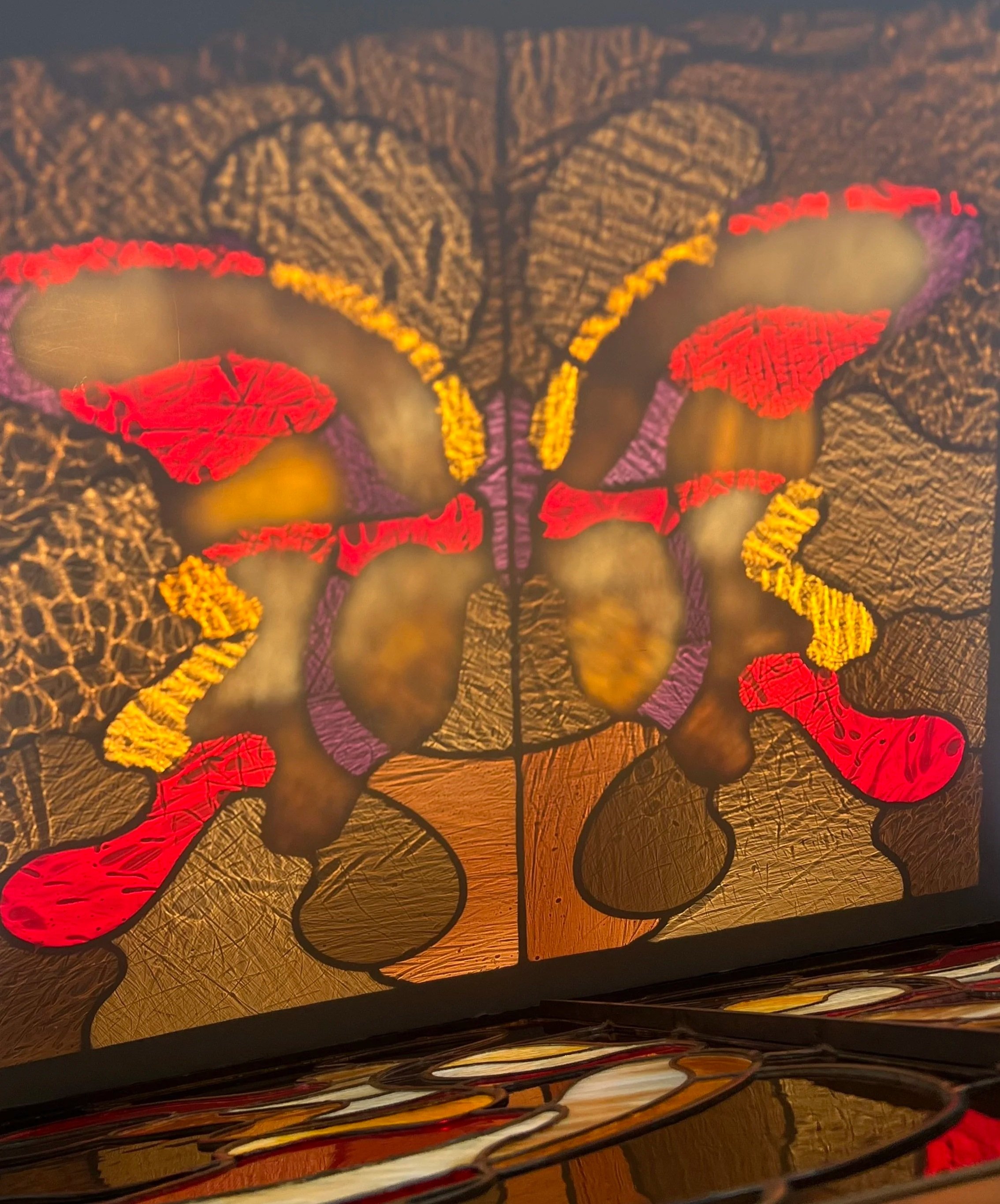 Close-up of a stained glass window with colorful butterfly and leaf designs, incorporating shades of red, purple, yellow, and black.