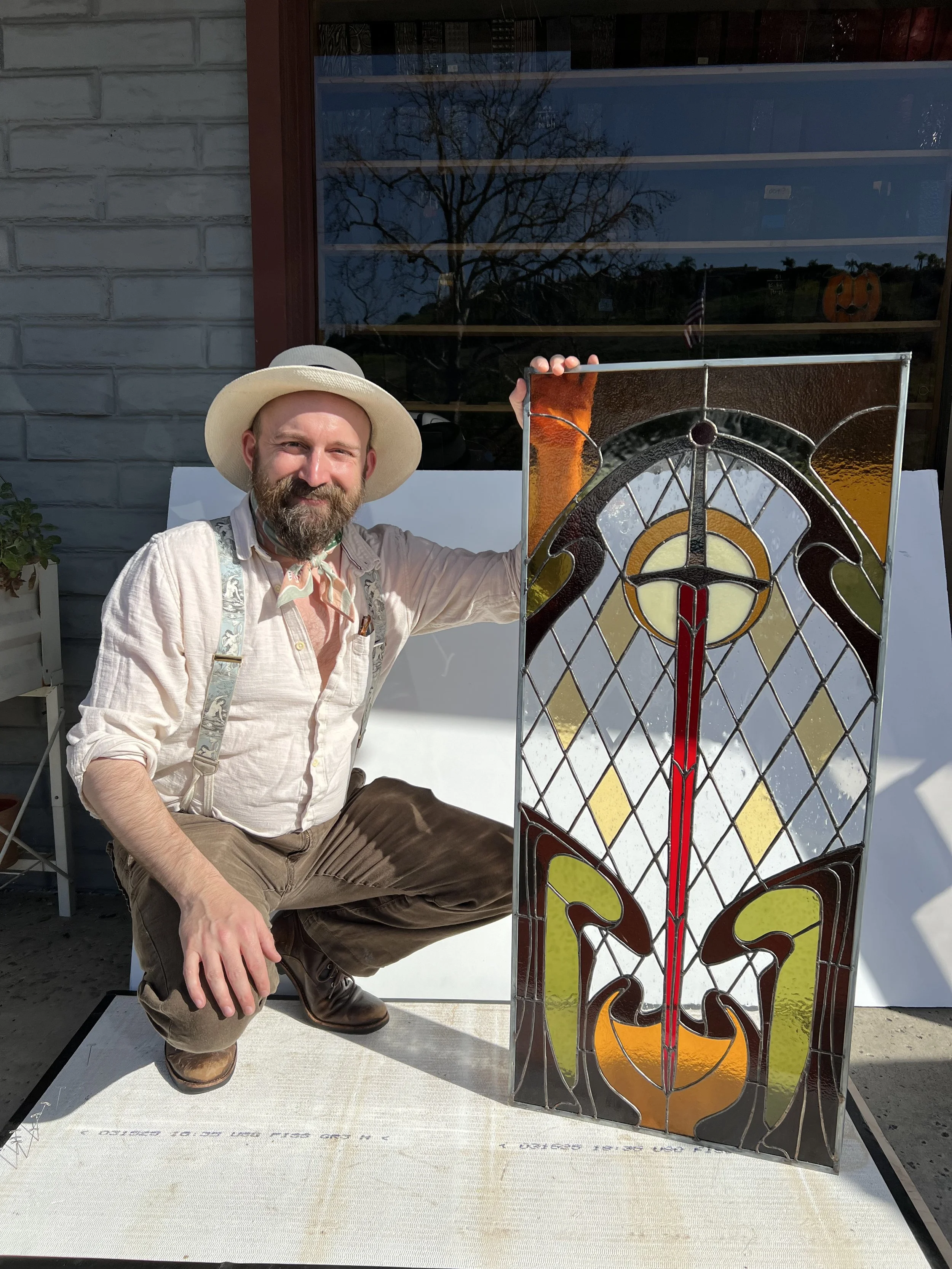 A man with a beard and wearing a wide-brimmed hat, beige shirt, and suspenders crouches next to a colorful stained glass butterfly art piece outdoors in front of a building window with a tree and sky reflection.