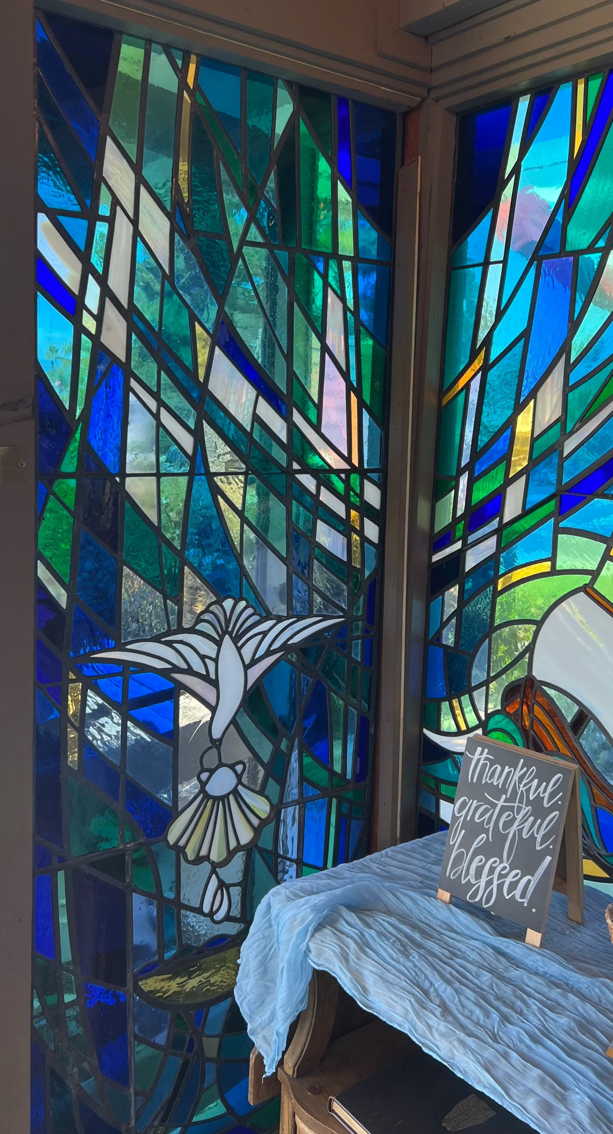 Colorful stained glass window featuring abstract shapes and a white dove.  A table with a white cloth in in front of the window, with a sign that says 'thankful, grateful, blessed'. Repair for Palisades Methodist Church in San Clemente