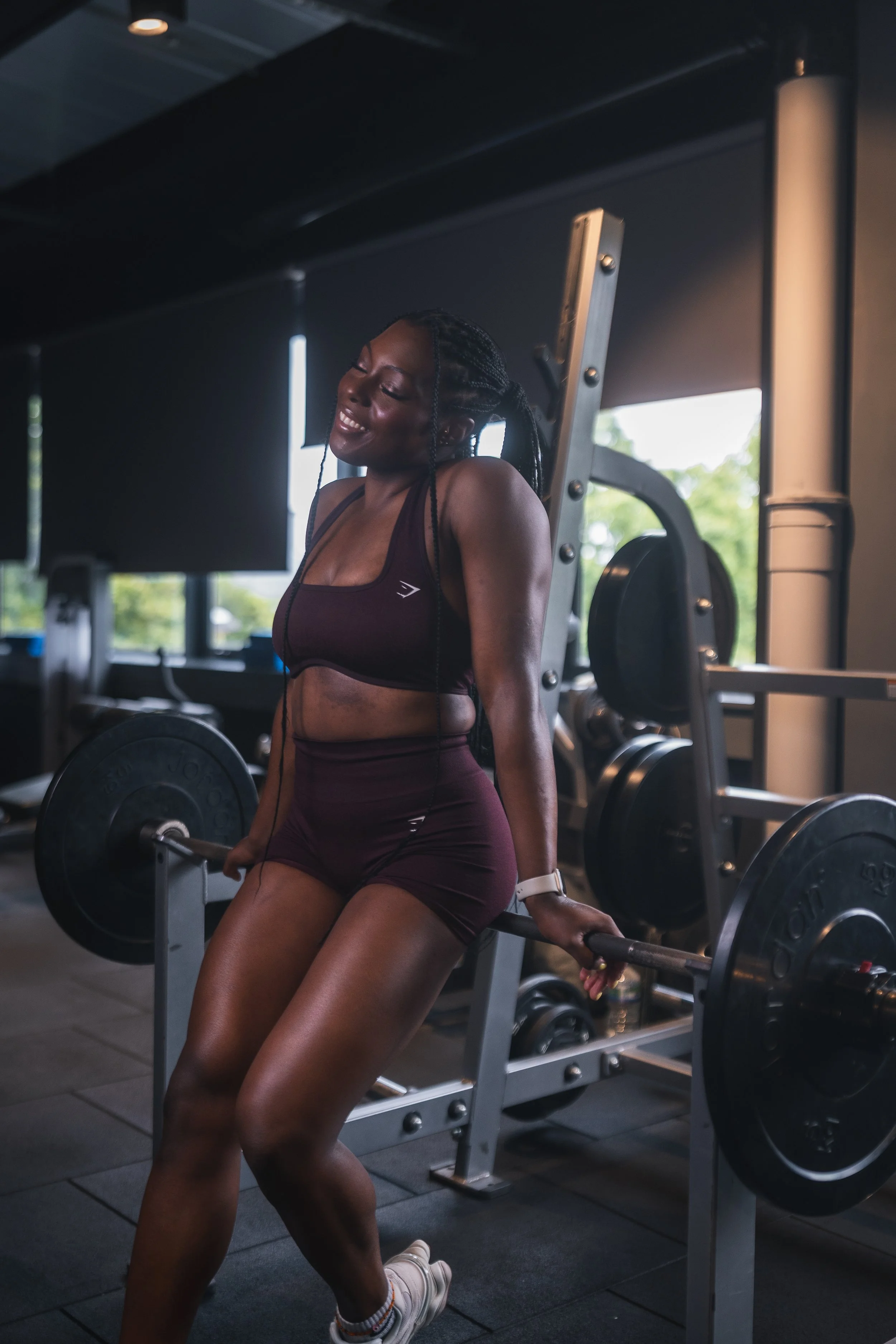 A woman in activewear smiling and leaning back on a barbell in a gym, with weightlifting equipment and windows in the background.