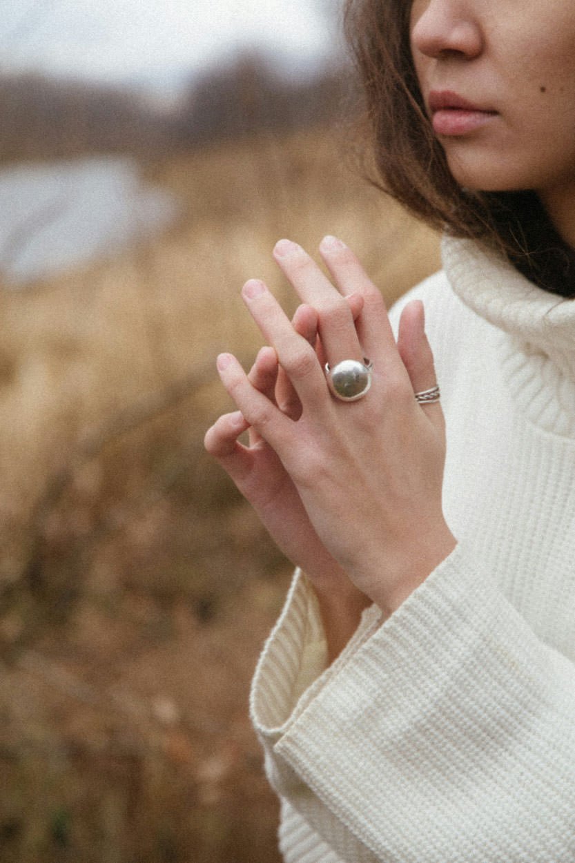 Close-up of a woman wearing a white sweater and two rings on her fingers, with hands pressed together, outdoors with blurred natural background.
