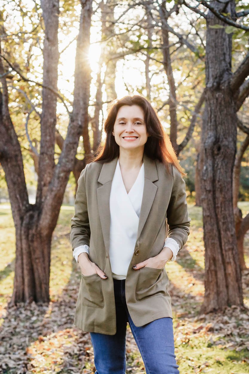 A woman standing outdoors in a park or wooded area during autumn, smiling at the camera with trees and fallen leaves in the background, backlit by sunlight.