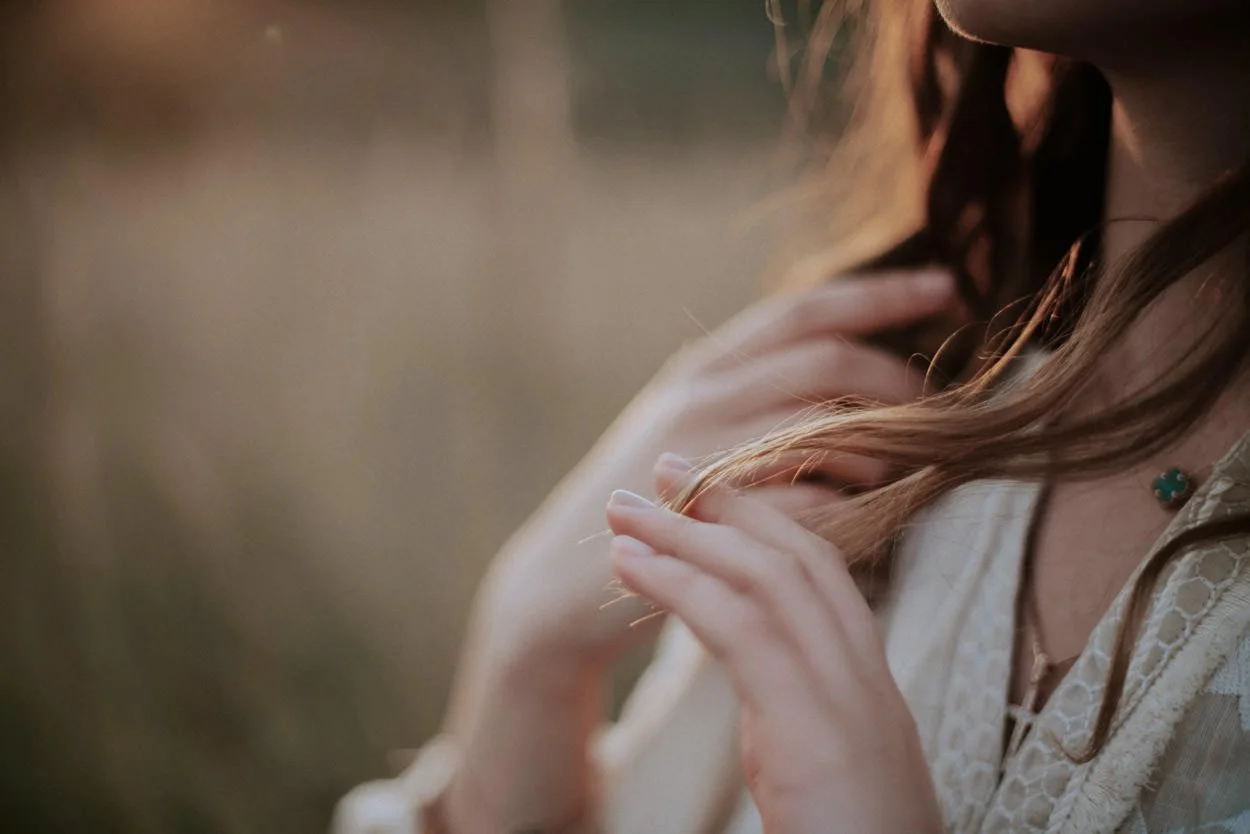 Close-up of a woman with long, light brown hair wearing a lace top and a turquoise necklace, gently touching her hair with her hands, outdoors during sunset.