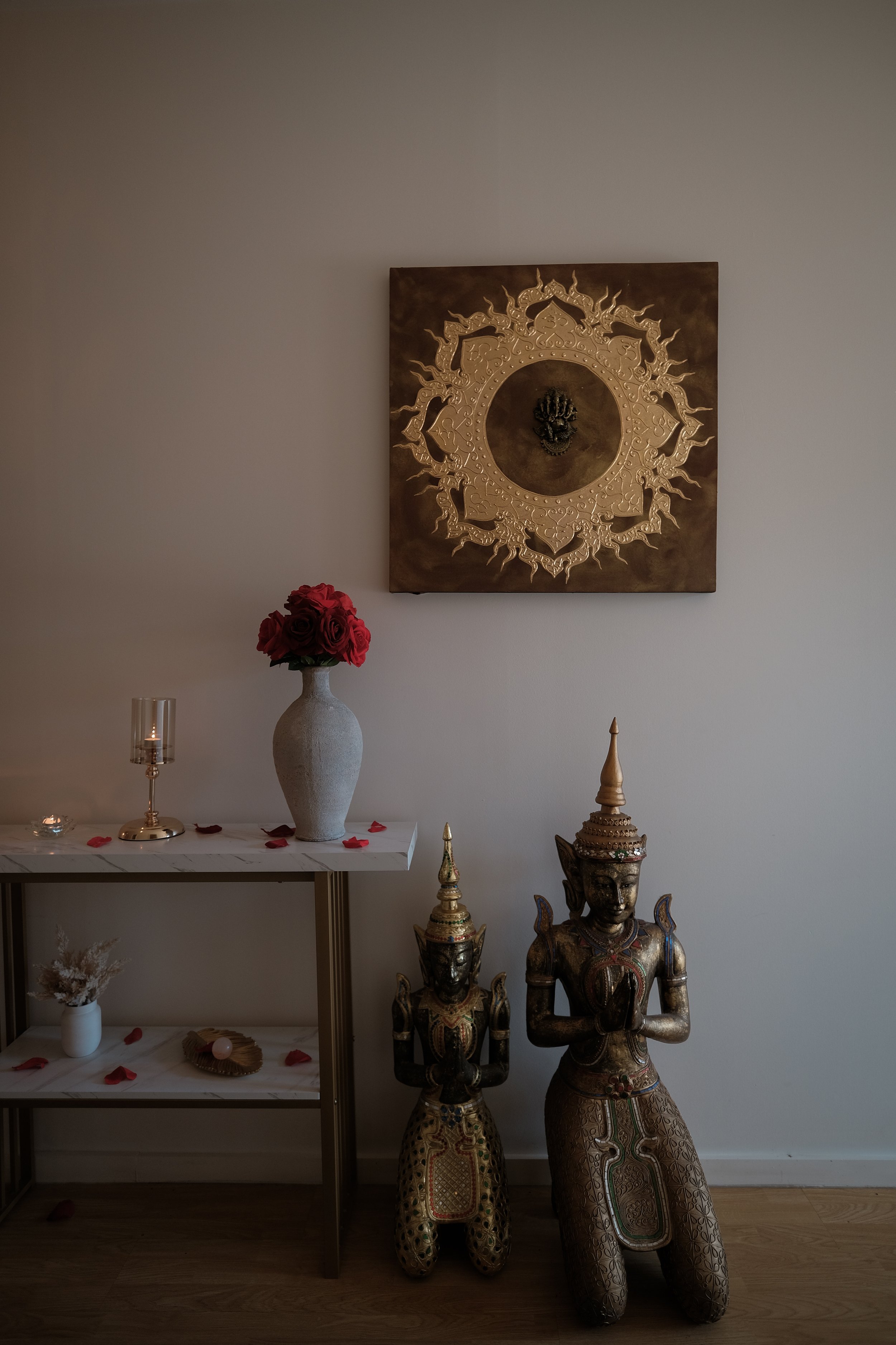 A beige wall with a square decorative artwork above two ornate Thai-style statues and a small table with a vase of red roses, a candle, and scattered rose petals.