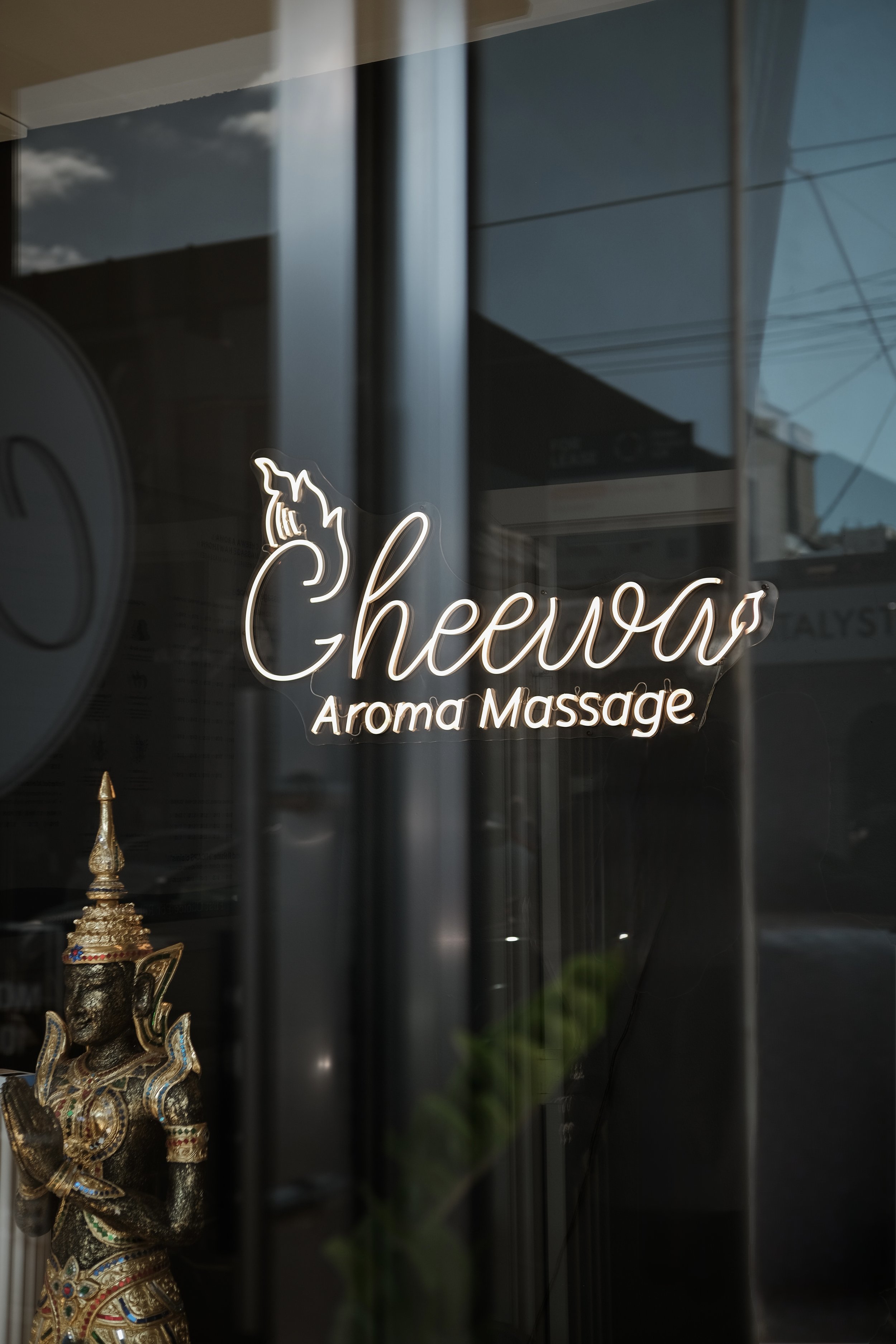 Neon sign reading 'Cheewa Aroma Remedial Thai Massage' with an illustration of a unicorn's head, inside a window reflecting buildings and a blue sky with clouds.