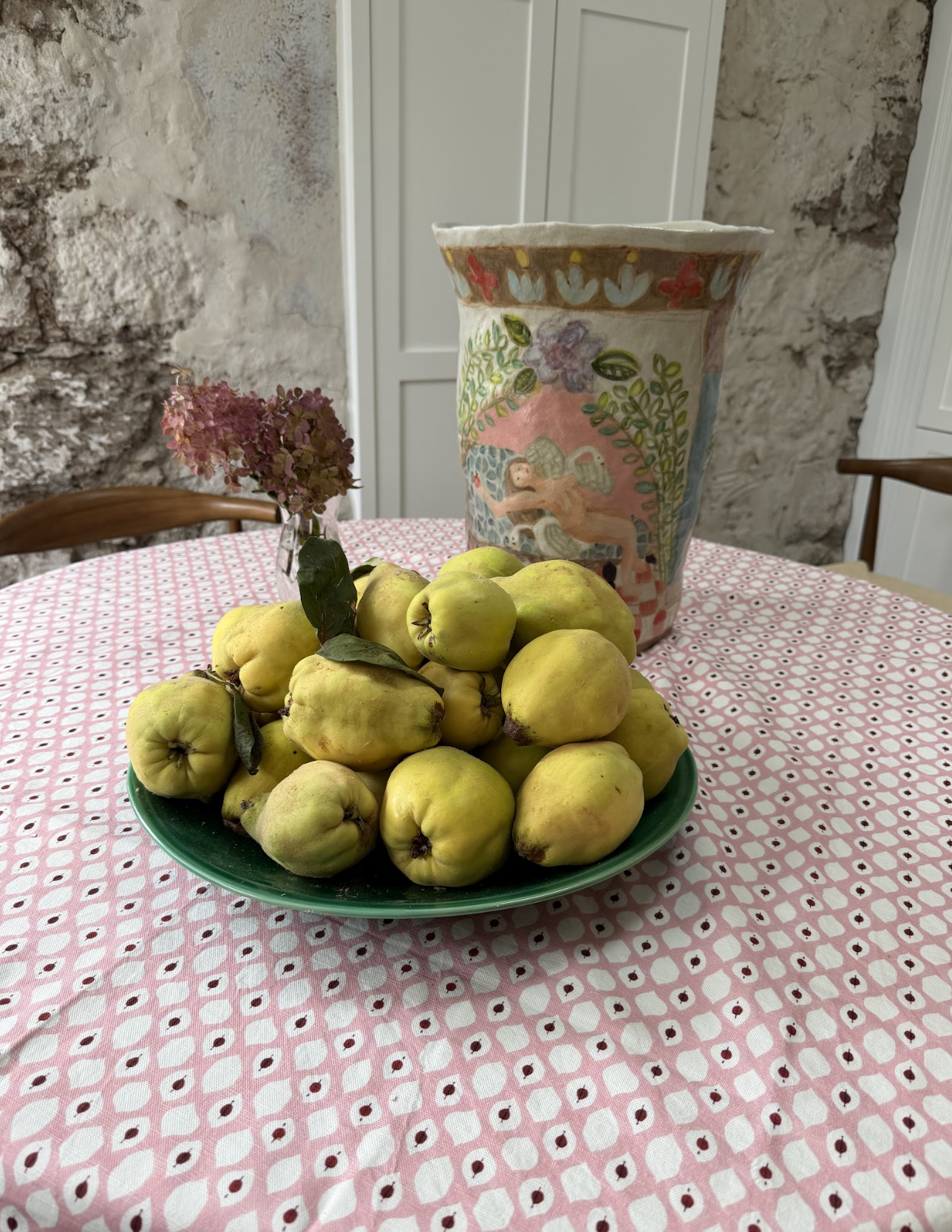 A plate of yellow quinces on a pink patterned tablecloth with a ceramic vase and a small vase with dried pink flowers in the background.