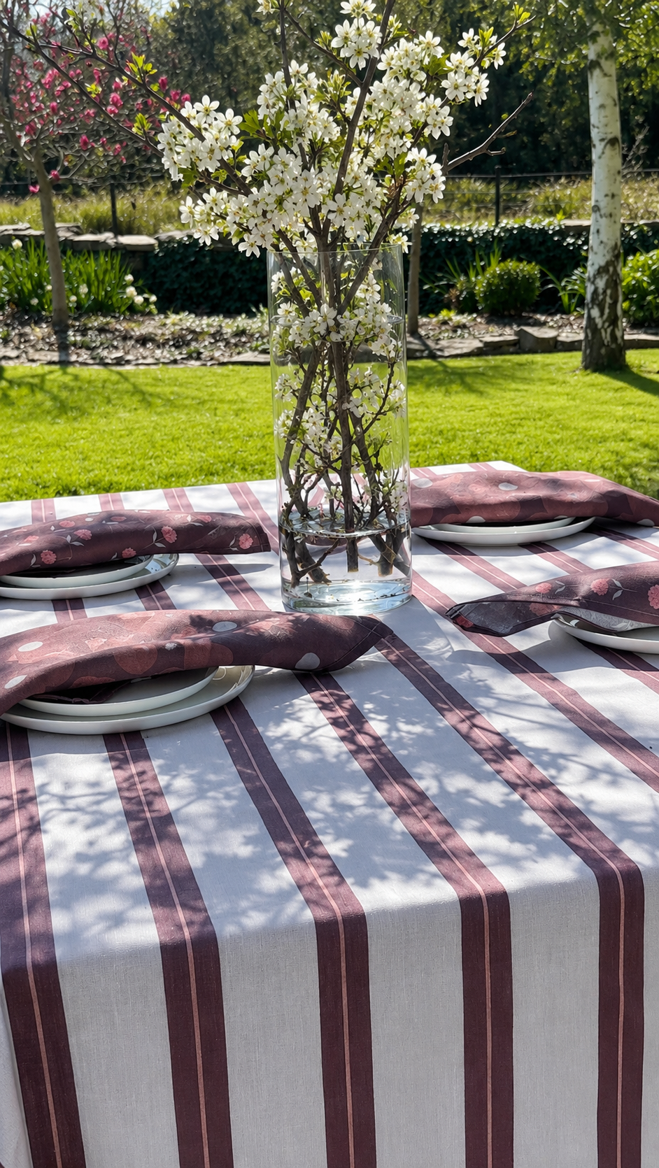 Outdoor table set for a meal with a vase of white blossoms as centerpiece, surrounded by plates and napkins, on a table with a striped tablecloth, in a garden with trees and green grass.