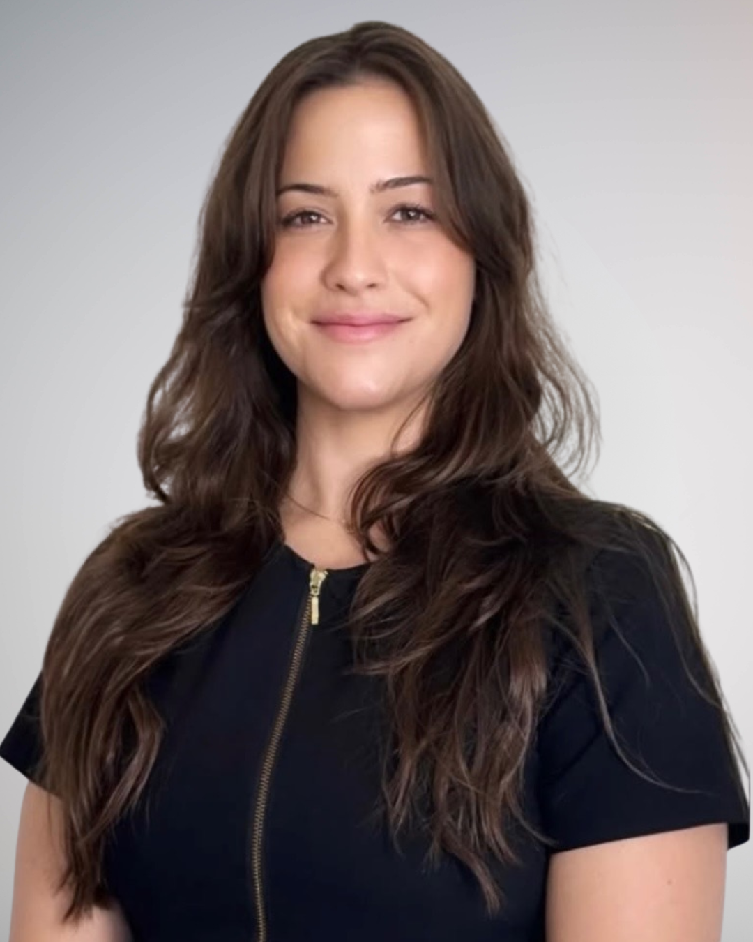 Portrait of a woman with long wavy brown hair, wearing a black top with a gold zipper, smiling against a neutral background.