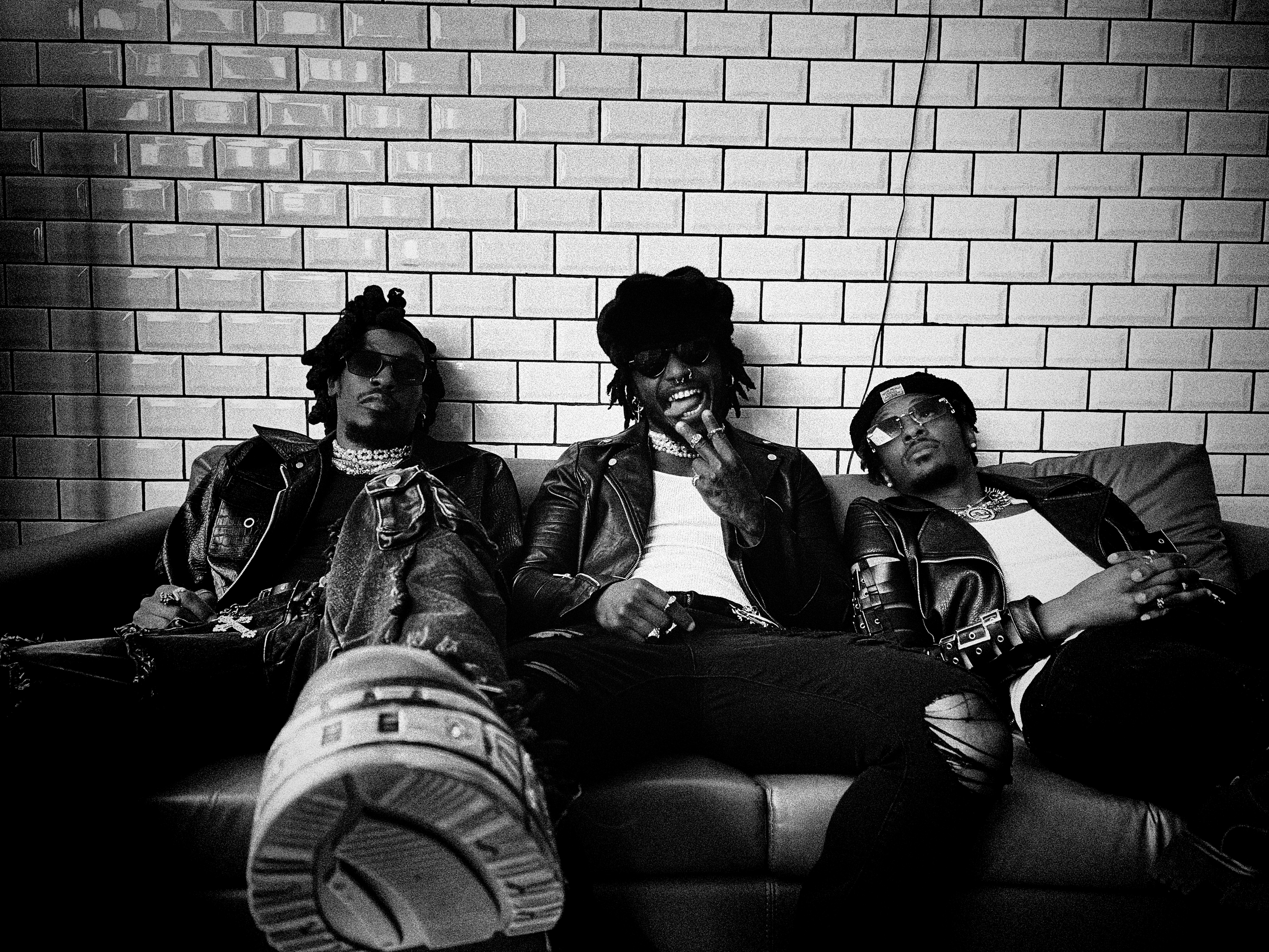 Hero The Band wearing sunglasses and leather jackets sitting against a brick wall, with their legs stretched out in front of them. The photo is in black and white.