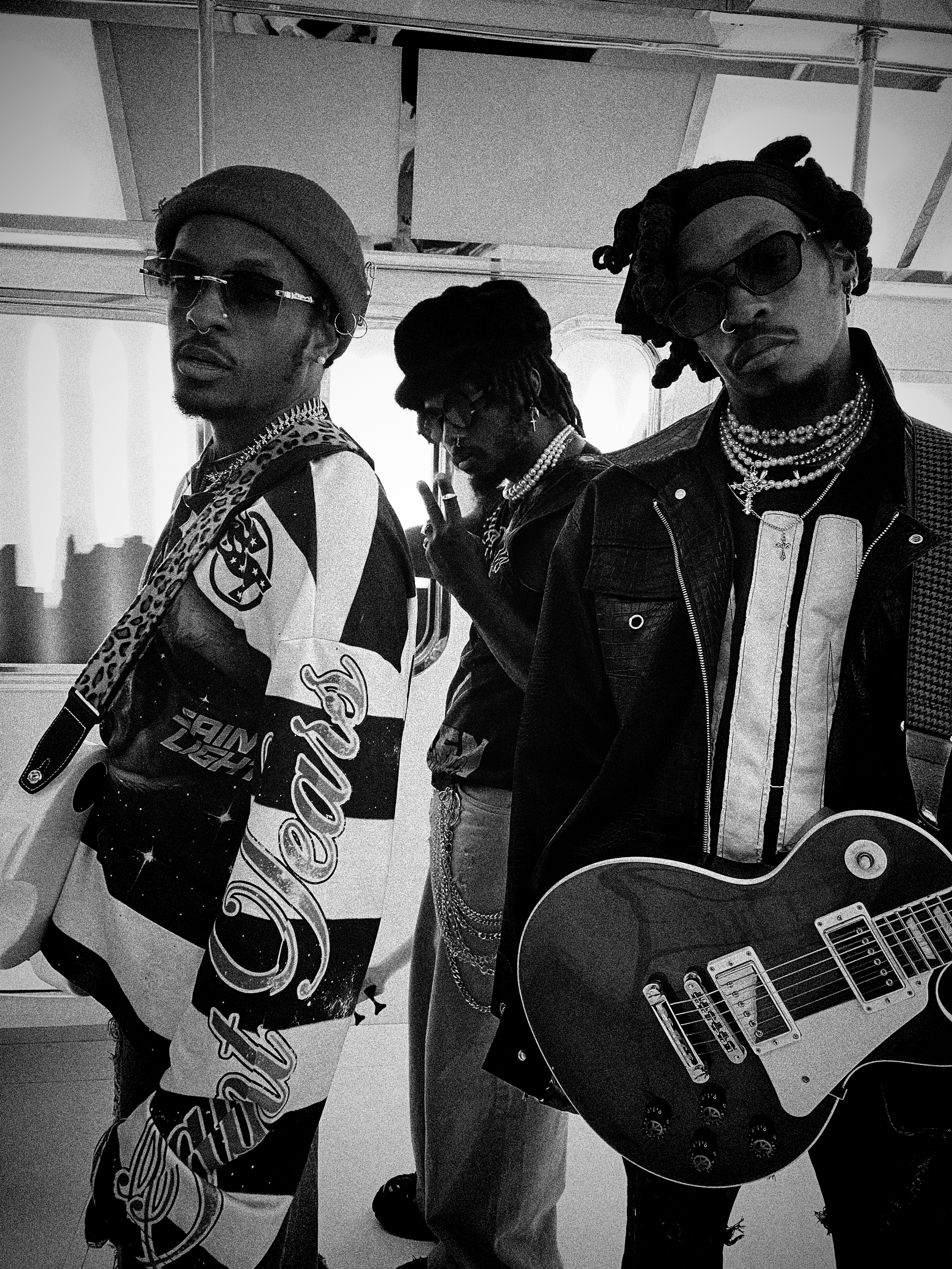 standing indoors. The person on the left wears sunglasses, a hat, a nose ring, and jewelry, and sports a patterned shirt. The person in the middle has dreadlocks, glasses, and is making a peace sign with their fingers. The person on the right wears sunglasses, a head covering, and layered necklaces, and holds an electric guitar.