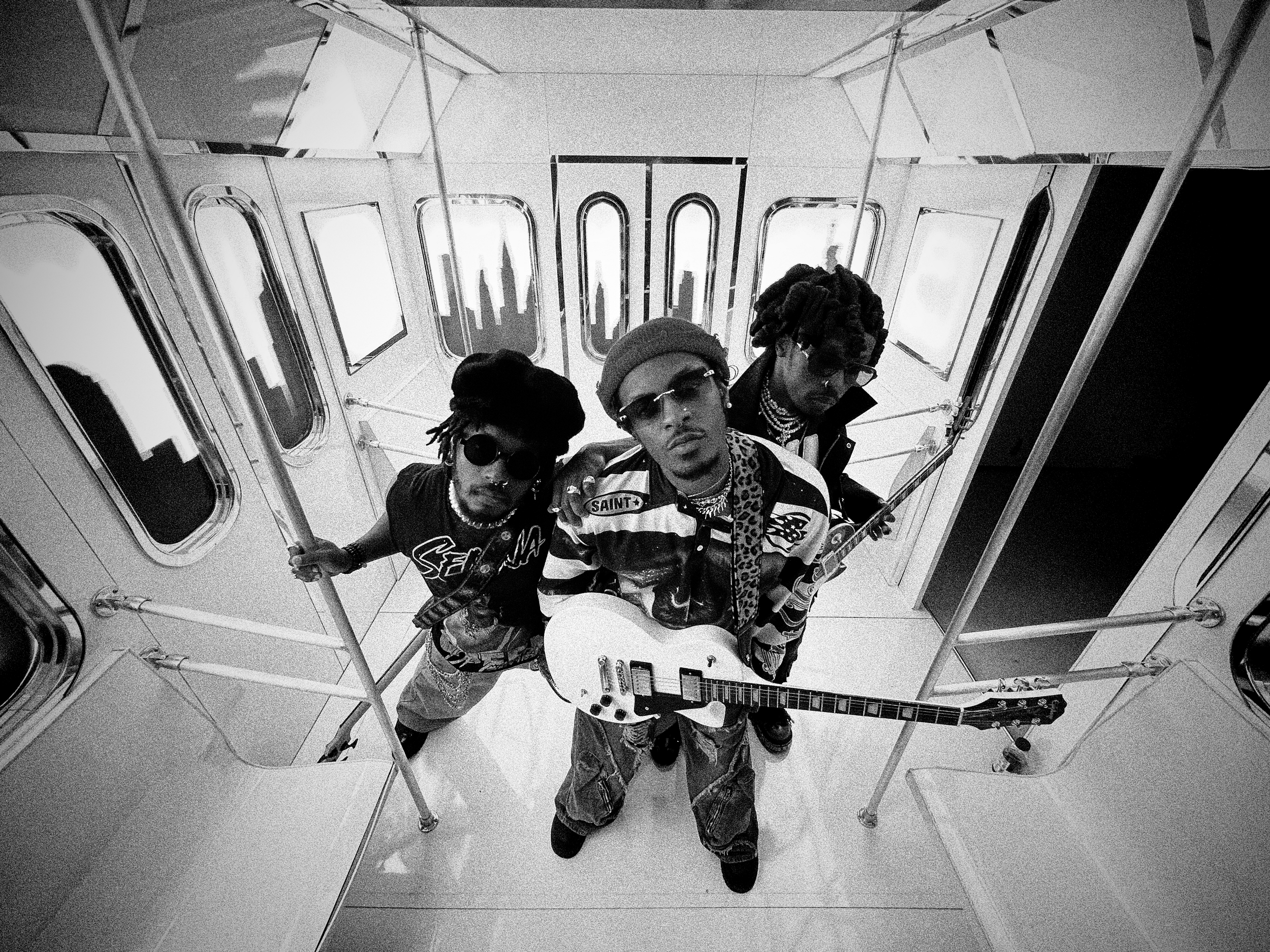Hero The Band standing on an escalator in a subway or train station, with three of them visible from the waist up and one laying on the ground, holding a guitar.
