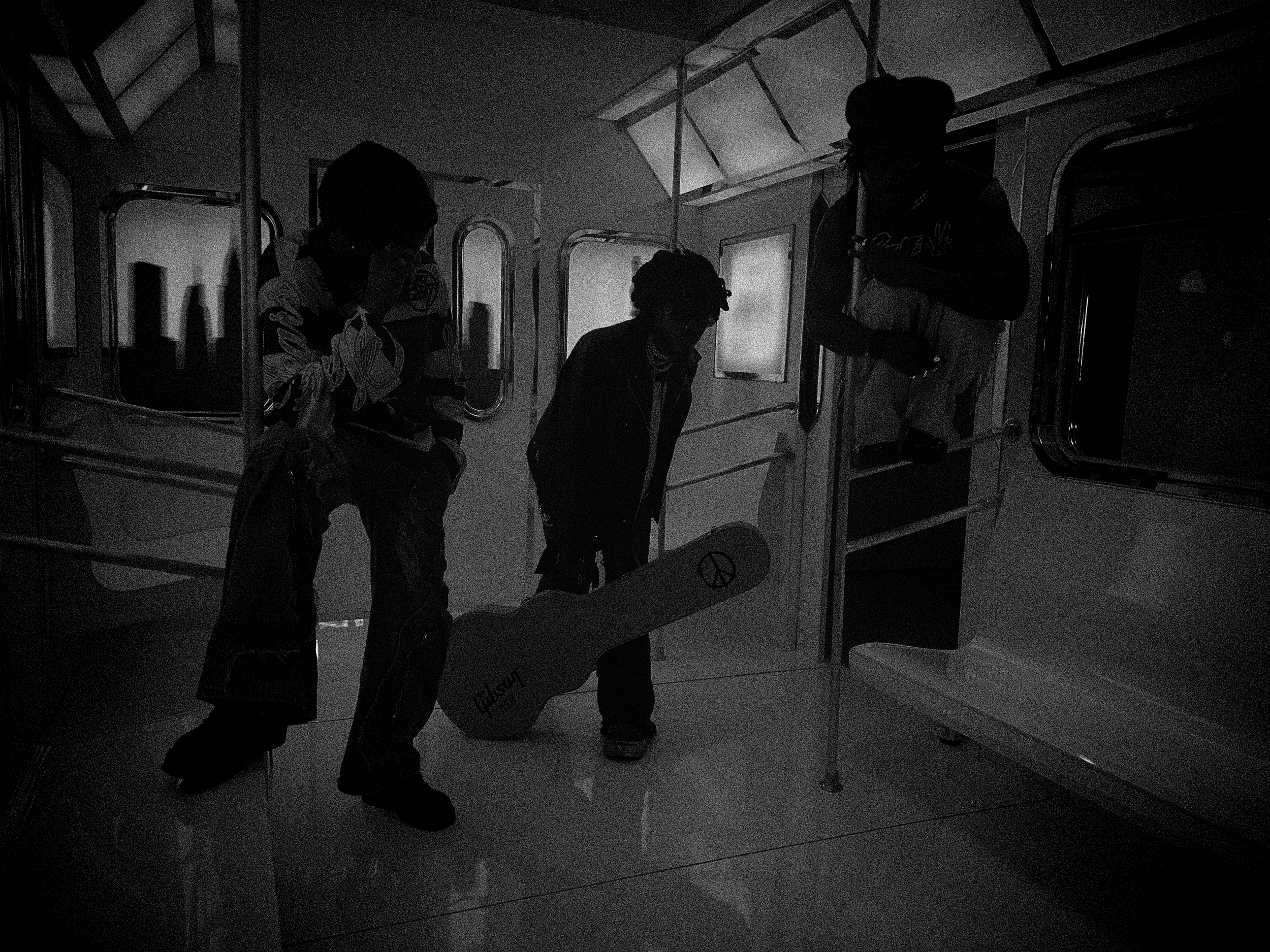 Hero The Band in a dark indoor setting, standing around a table with guitar cases, with a window and framed picture on the wall in the background.