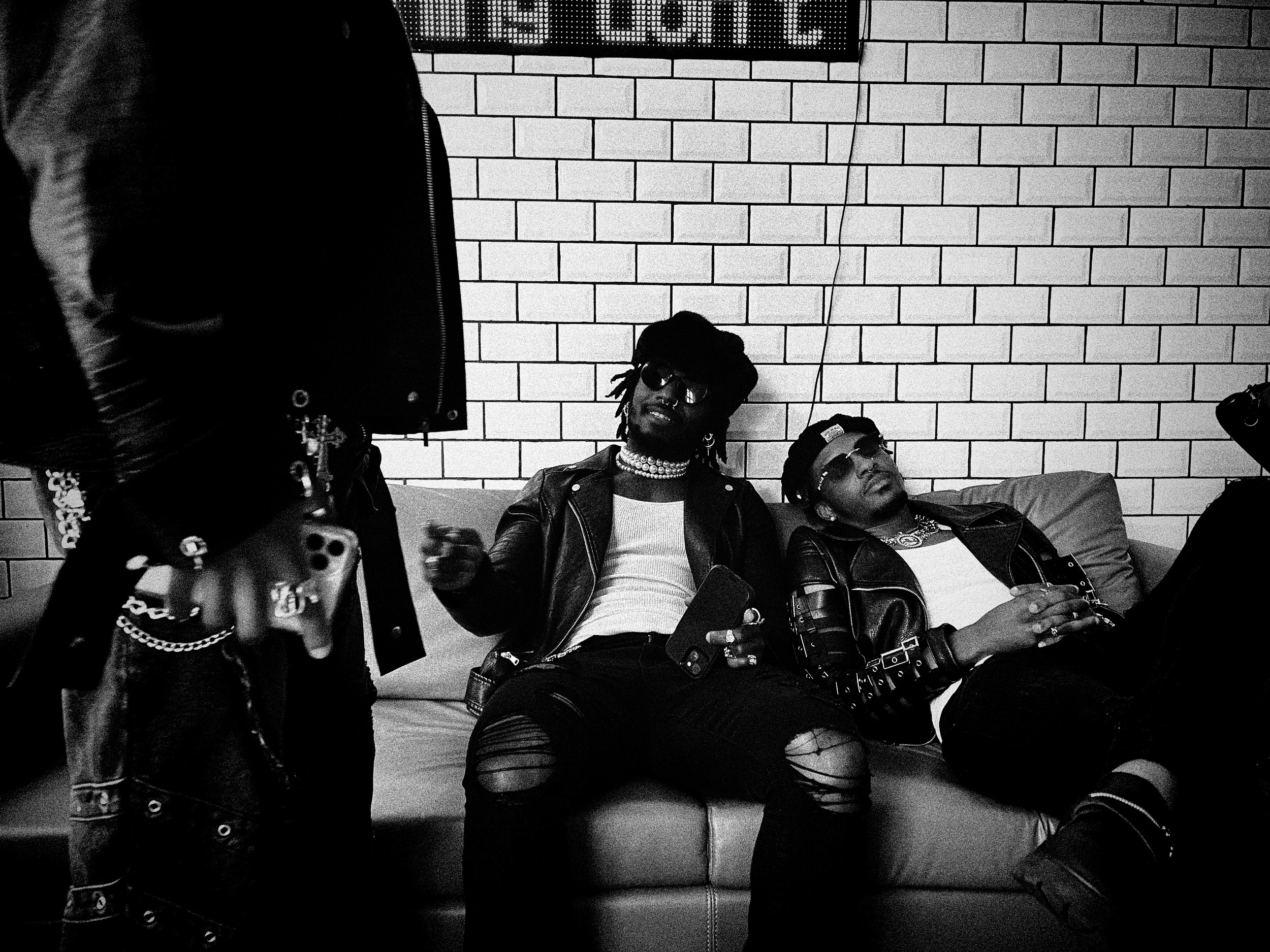 People sitting and standing against a brick wall, some wearing sunglasses and jewelry, in a black-and-white photo.