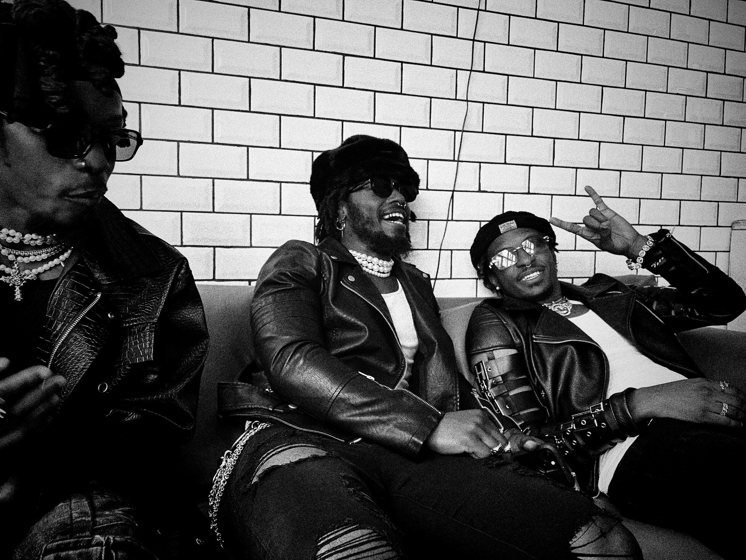 Group of four people sitting against a brick wall, all wearing leather jackets and sunglasses, smiling, with one making a peace sign.