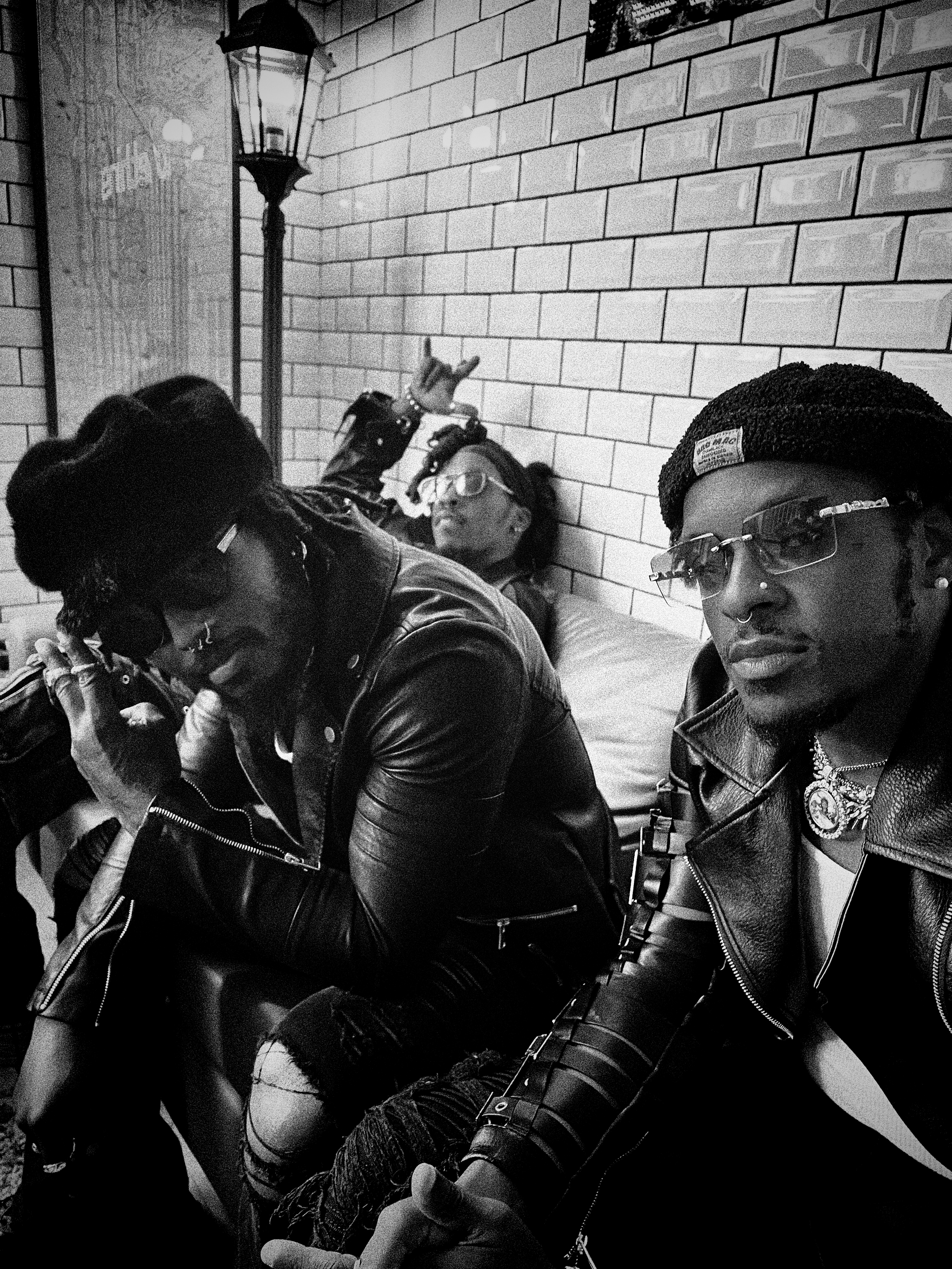 sitting against a brick wall, all wearing jackets and sunglasses, with hairstyles and accessories typical of a punk or alternative fashion style. One person is making a rock hand gesture, and another is showing their phone in an indoor setting with a wall-mounted lamp.