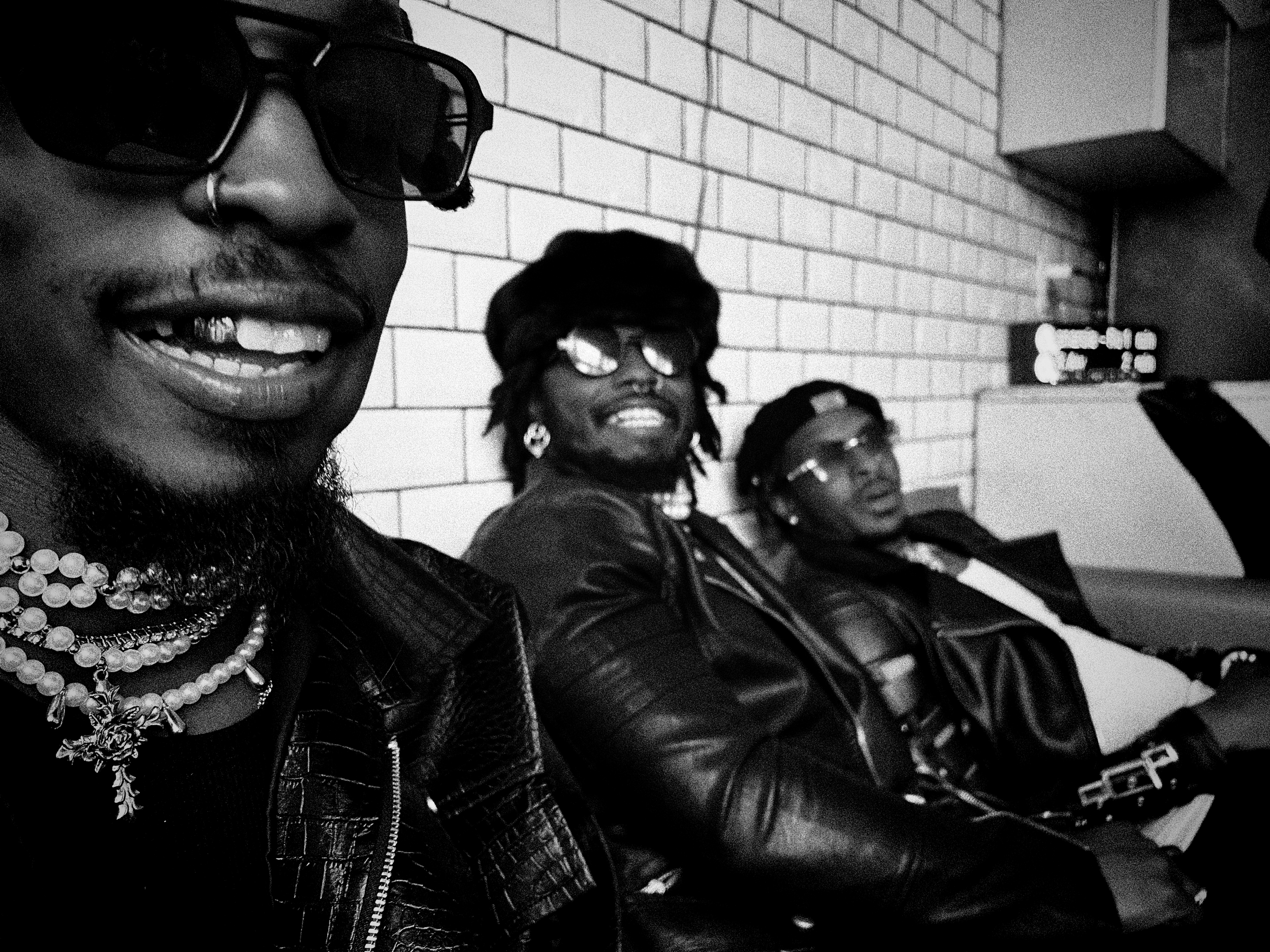 Hero The Band, Close-up wearing sunglasses, jewelry, and a leather jacket, sitting with other people against a brick wall in black and white.