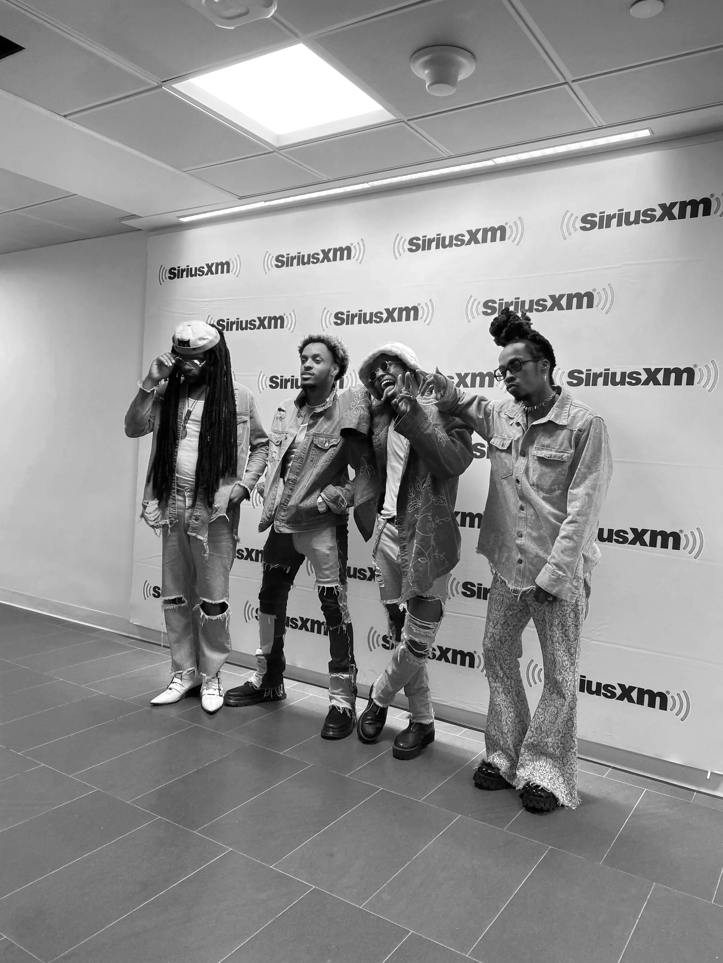 Hero The Band standing in front of a step and repeat banner with the SiriusXM logo, posing for a photo at what appears to be a promotional or media event, with casual style and unique fashion choices.