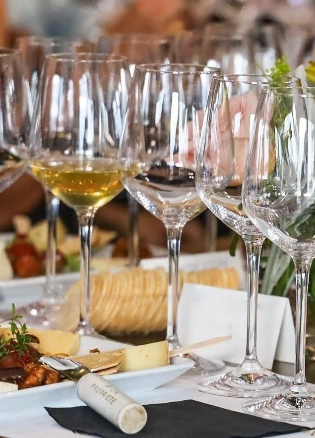 Multiple empty and partially filled wine glasses on a table set for a gathering with cheese, bread, and charcuterie in the background.